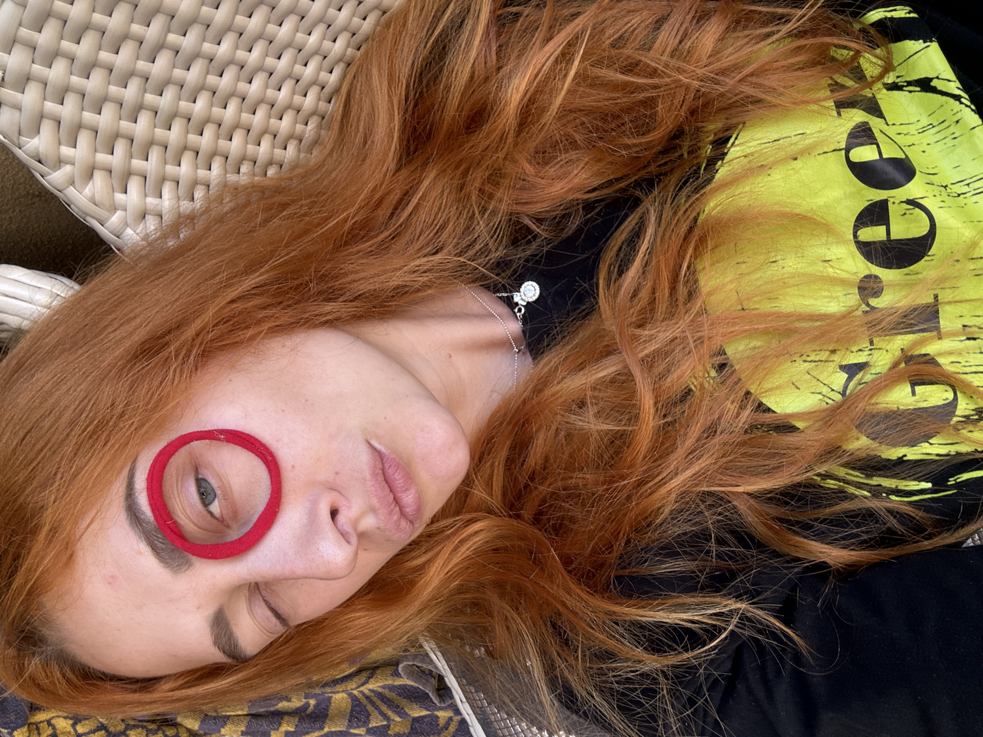 A red-haired woman reclines on a wicker chair, a red hair tie encircling her left eye. She has fair skin, a silver necklace, and is winking. Her head rests on a patterned cushion. She wears a black shirt with a neon yellow graphic on her right side. The scene is outdoors, possibly in Budva, Montenegro, given the image's origin.