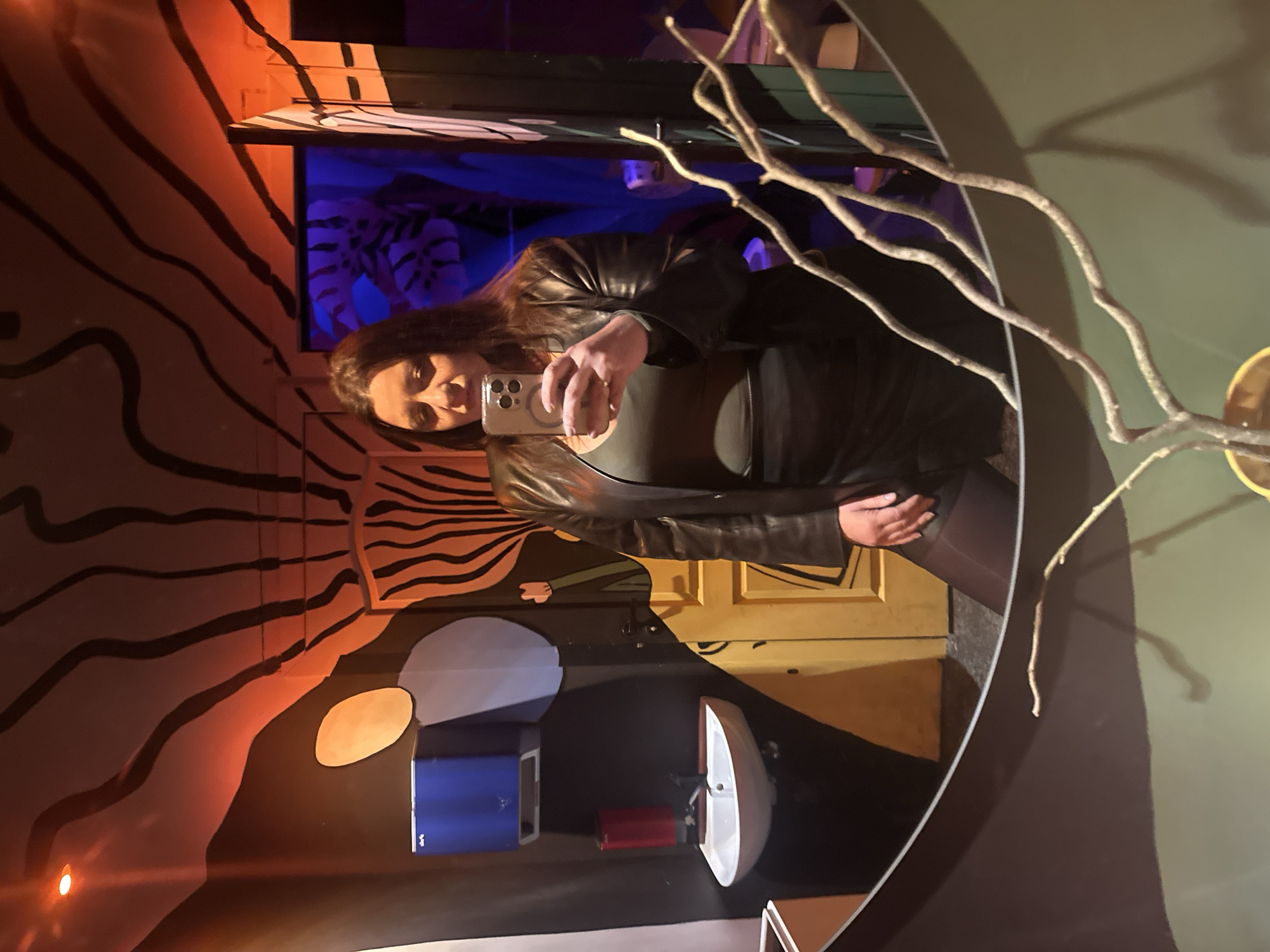 A woman wearing a black leather jacket is taking a selfie in a bathroom with decorative mirrors. The bathroom has orange walls with a black zebra-stripe pattern, light fixtures in the shape of circles, a blue cabinet, a red container, and a white sink. There is also a decorative tree branch emerging from the circular mirror on the right. A painting or mural behind the mirror depicts foliage in blue. The location appears to be a public restroom, potentially in an establishment such as a bar or restaurant, based on the artistic decorations. The specific location in Riga, Latvia, cannot be confirmed from the image alone.