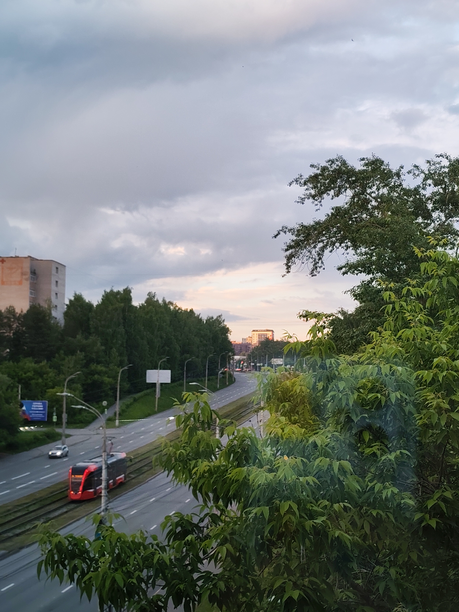 The image depicts a view from an elevated position, likely a building, overlooking a street in Izhevsk, Russia. The sky is overcast with a hint of sunset colors near the horizon. In the midground, a multi-lane road is visible, with a tram line running along its center. A red and grey tram is currently on the tracks, heading in the direction of the viewer, with a silver car ahead of it in the roadway. Trees line the side of the road and foreground. In the background, several buildings can be seen, including a multi-story residential building and other structures further away. The time of day appears to be evening or late afternoon.