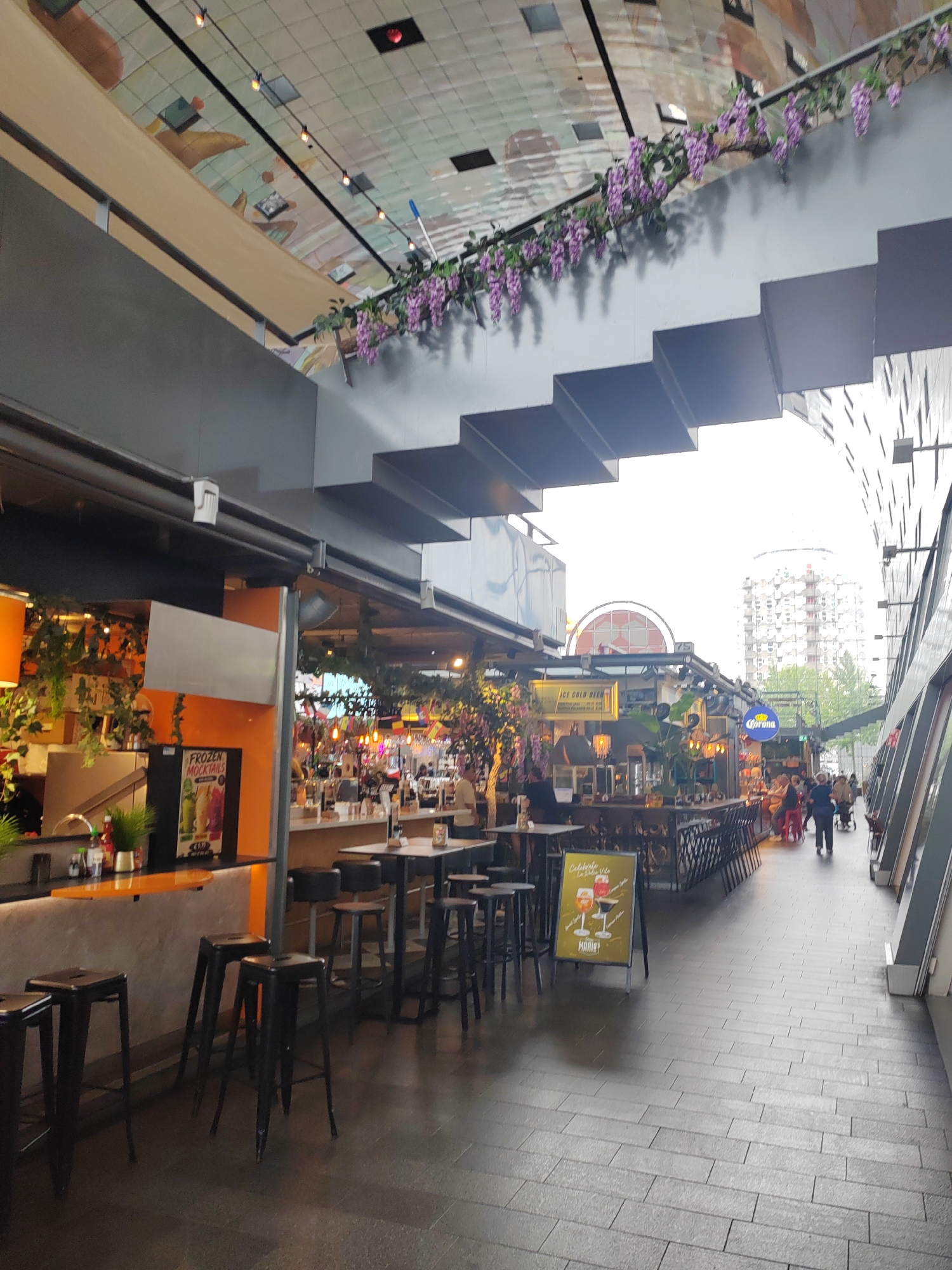 This image captures a vibrant indoor market or food hall in Rotterdam, Netherlands. The scene depicts a bustling commercial space with various food and drink stalls lining a wide walkway. In the foreground, several tall, black stools are arranged around counter-height tables, suggesting a seating area for patrons. Further down the walkway, patrons are visible at various bars and tables, indicating an active social and dining environment.
The architecture of the space is notable, with a creatively decorated ceiling featuring colorful artwork and an interesting stepped design along the upper levels. Artificial purple flowers hang from a railing, adding a touch of natural decoration. In the background, glimpses of buildings, including a distinctive round-roofed structure and a modern high-rise, provide context for the urban setting.
Several signs are visible, including one for "FROZEN MOCKTAILS" and another yellow sign advertising "ICE COLD BEER" with a Corona logo. A freestanding yellow sign in the walkway displays an image of two drinks and the text "Celebrate La Dolce Vita MARIO!".
The lighting suggests it might be daytime, possibly late afternoon or early evening, given the mix of natural and artificial light and the indoor setting. The overall atmosphere appears lively and inviting, typical of a popular food and social hub. The presence of people engaged in dining and socializing suggests an ongoing daily activity.