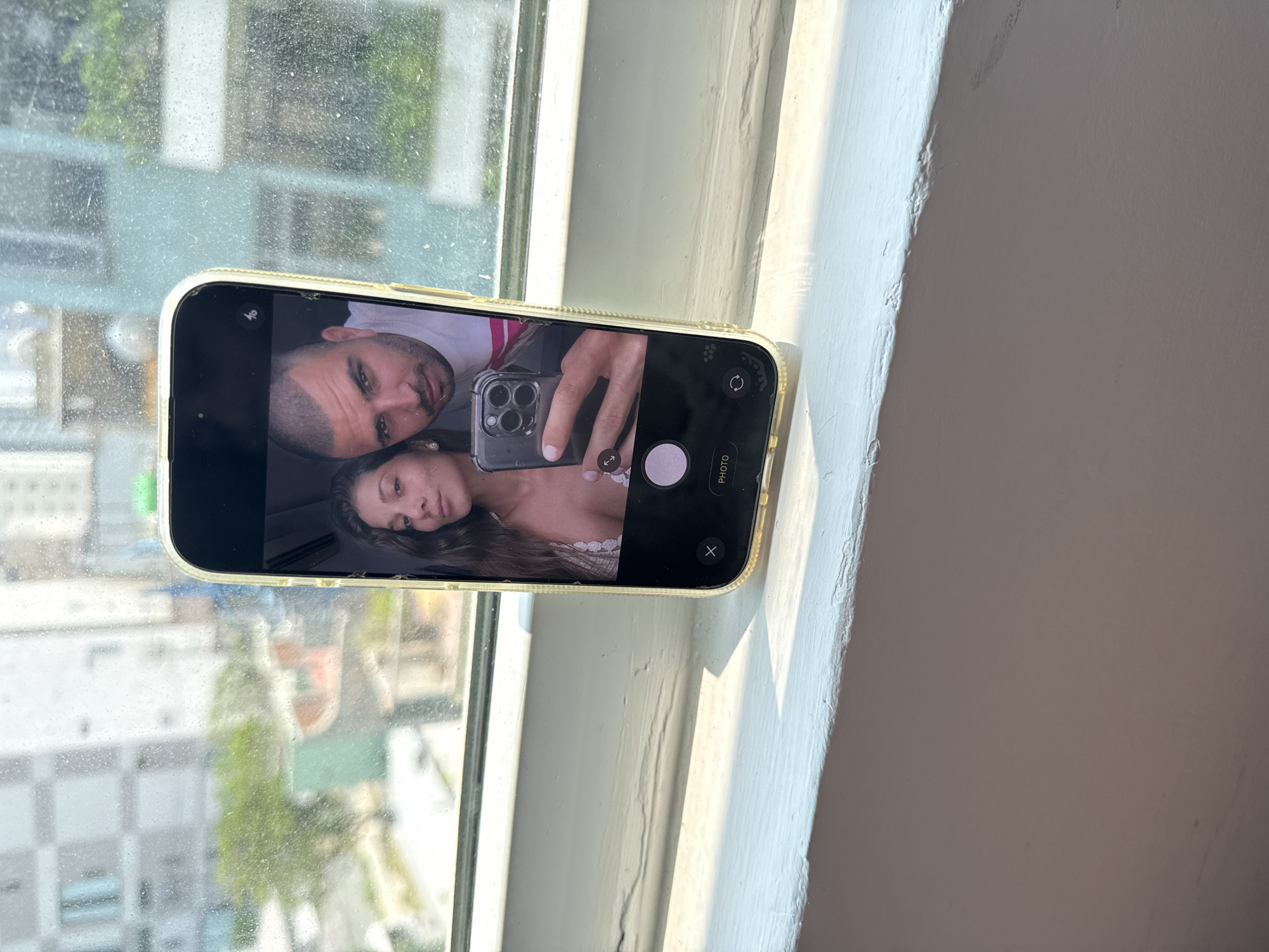 A smartphone displaying a selfie of a man and a woman is resting on a windowsill. The phone is in the camera app. The man has short hair and is wearing a light-colored shirt. The woman has long brown hair. The windowsill is white and has paint flaws. Outside the window, buildings and trees are visible, likely in a city. The city is Đà Nẵng, Vietnam. The photo has natural light. The camera of the smartphone on the windowsill has three lenses.