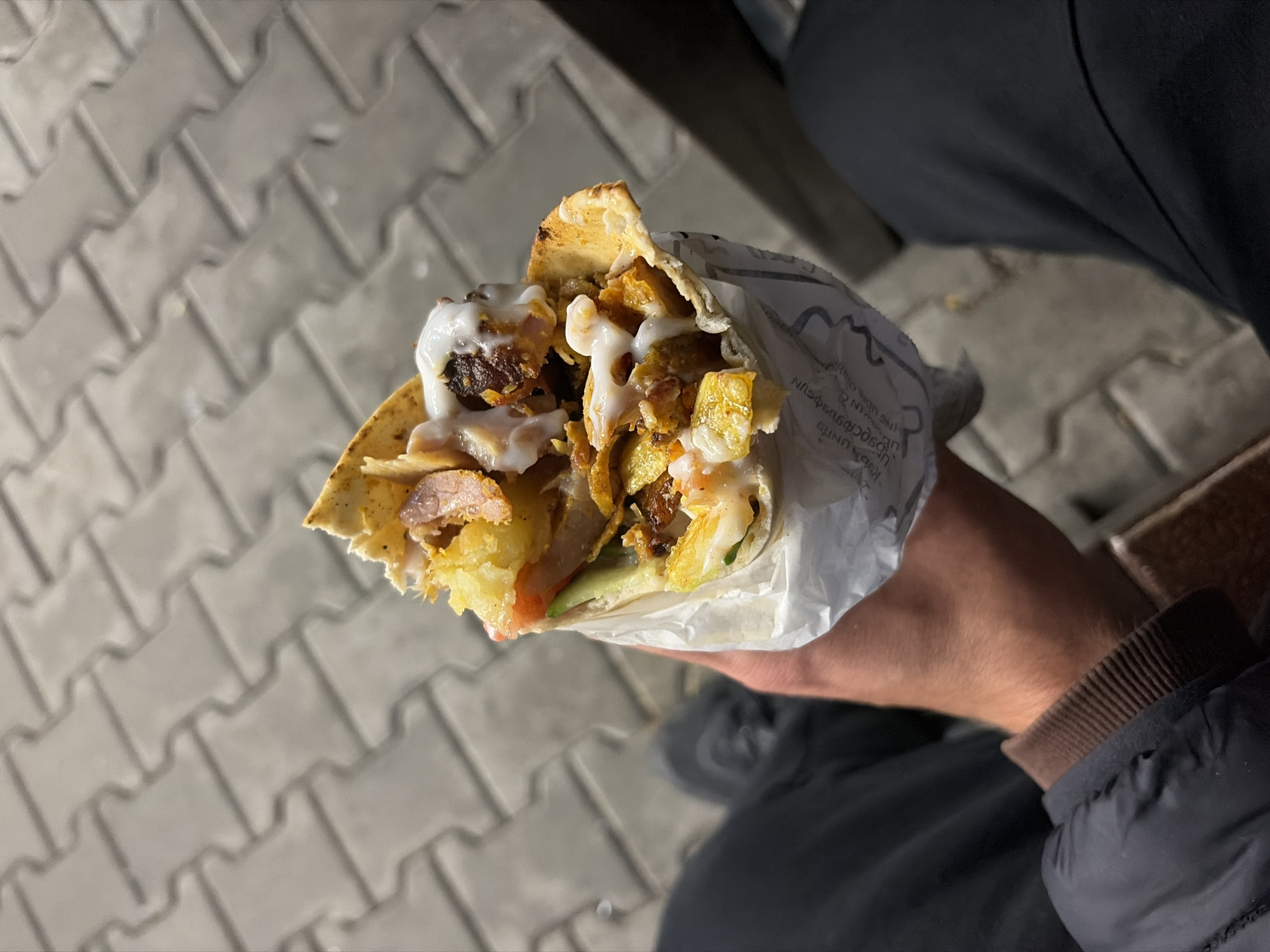 A first-person perspective photograph depicts a hand holding a shawarma wrap. The wrap consists of meat, vegetables, sauce, and possibly fried potatoes within a thin flatbread. The wrap is partially enclosed in white paper. The background includes a gray interlocking brick sidewalk, and the person wearing dark-colored clothing. The location is purportedly in Veliko Tarnovo, Bulgaria, although there is no confirmation of this in the image.