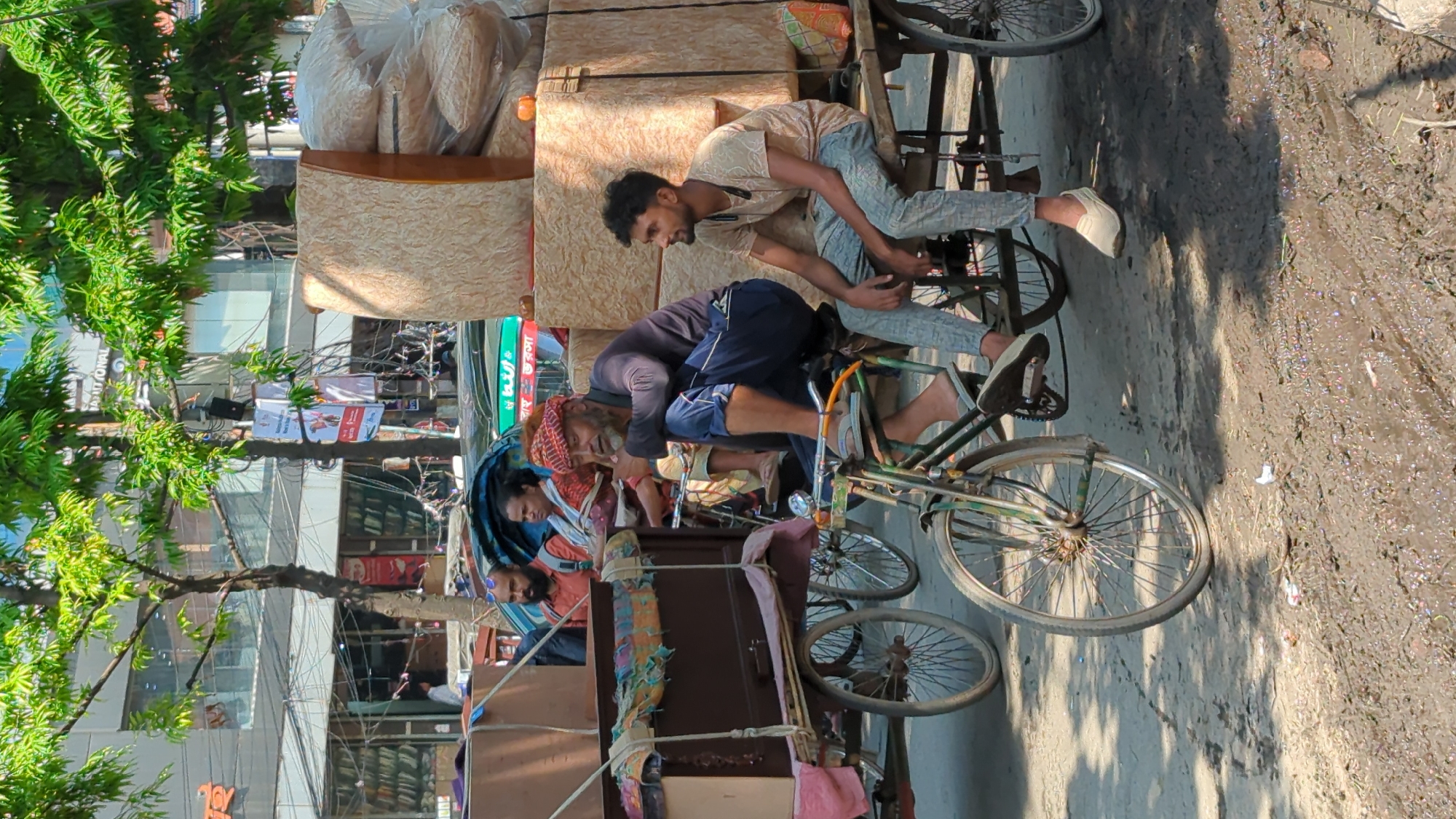 In Dhaka, Bangladesh, a man with a red head covering is seated on the back of a bicycle rickshaw, which is heavily loaded with furniture and other items. A younger man in a light-colored shirt is also seated on the rickshaw, appearing to assist or observe. The rickshaw is on a street with a muddy, wet surface. Buildings and trees are visible in the background, indicating an urban environment. Power lines crisscross the sky.