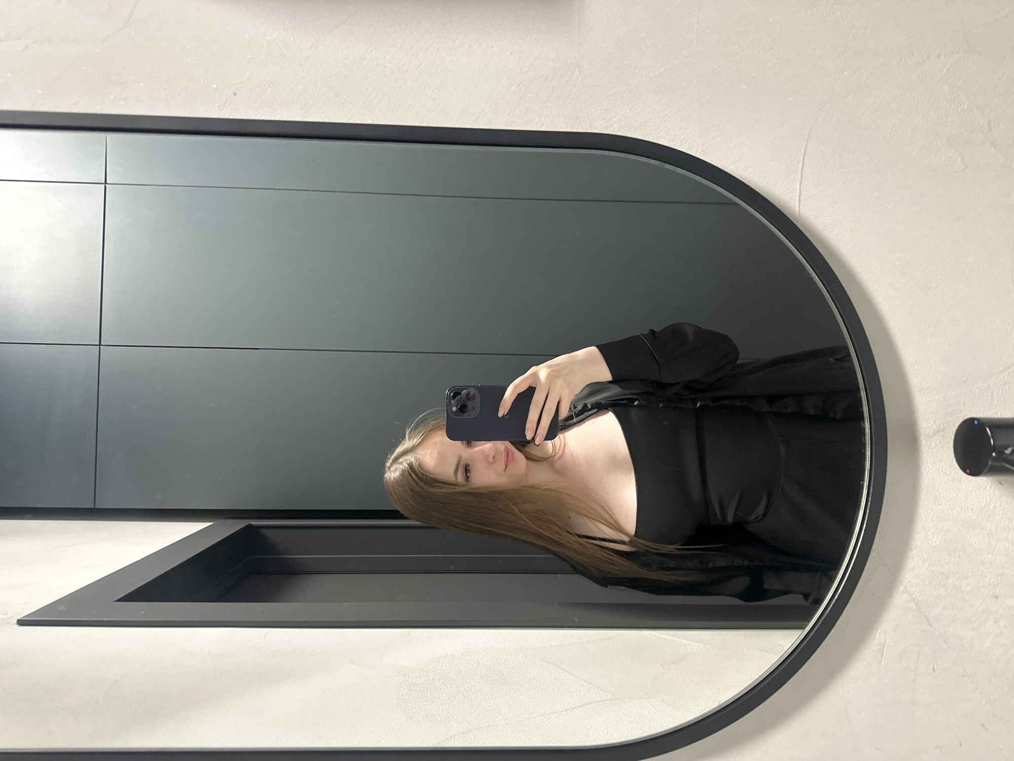 A young woman with long, light brown hair is taking a selfie in a mirror. She is wearing a black camisole and a black open-front jacket. She is holding a black smartphone in her right hand, and her left hand is resting on the phone. The mirror is oval-shaped with a black frame and is reflecting a grey, textured wall and a grey door or cabinet. The woman is looking at the camera with a slight smile. The environment appears to be indoors, possibly a bathroom or a dressing room.