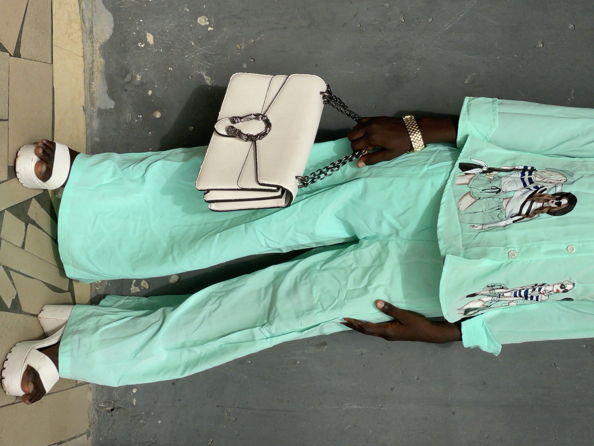 A person lies on the ground in Badagry, Nigeria. The person is wearing a light green two-piece outfit with drawings of people on the shirt. The person is also wearing white platform sandals and a gold watch. A white handbag with a silver chain sits on the person's midsection. The ground is a mix of concrete and tile.