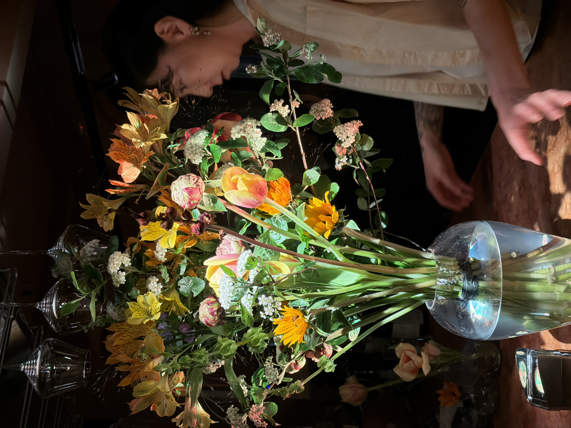 A floral arrangement in a glass vase is on a dark, reflective surface. The arrangement includes sunflowers, roses, alstroemeria, tulips, and other flowers, along with greenery. To the left of the bouquet are three wine glasses. A person with dark hair is partially visible above the bouquet, wearing a light-colored top and dark pants, with their hand extended towards the surface. The setting is dim and indoors. The location is unclear, and there is no discernible evidence to suggest it is Moscow, Russia.