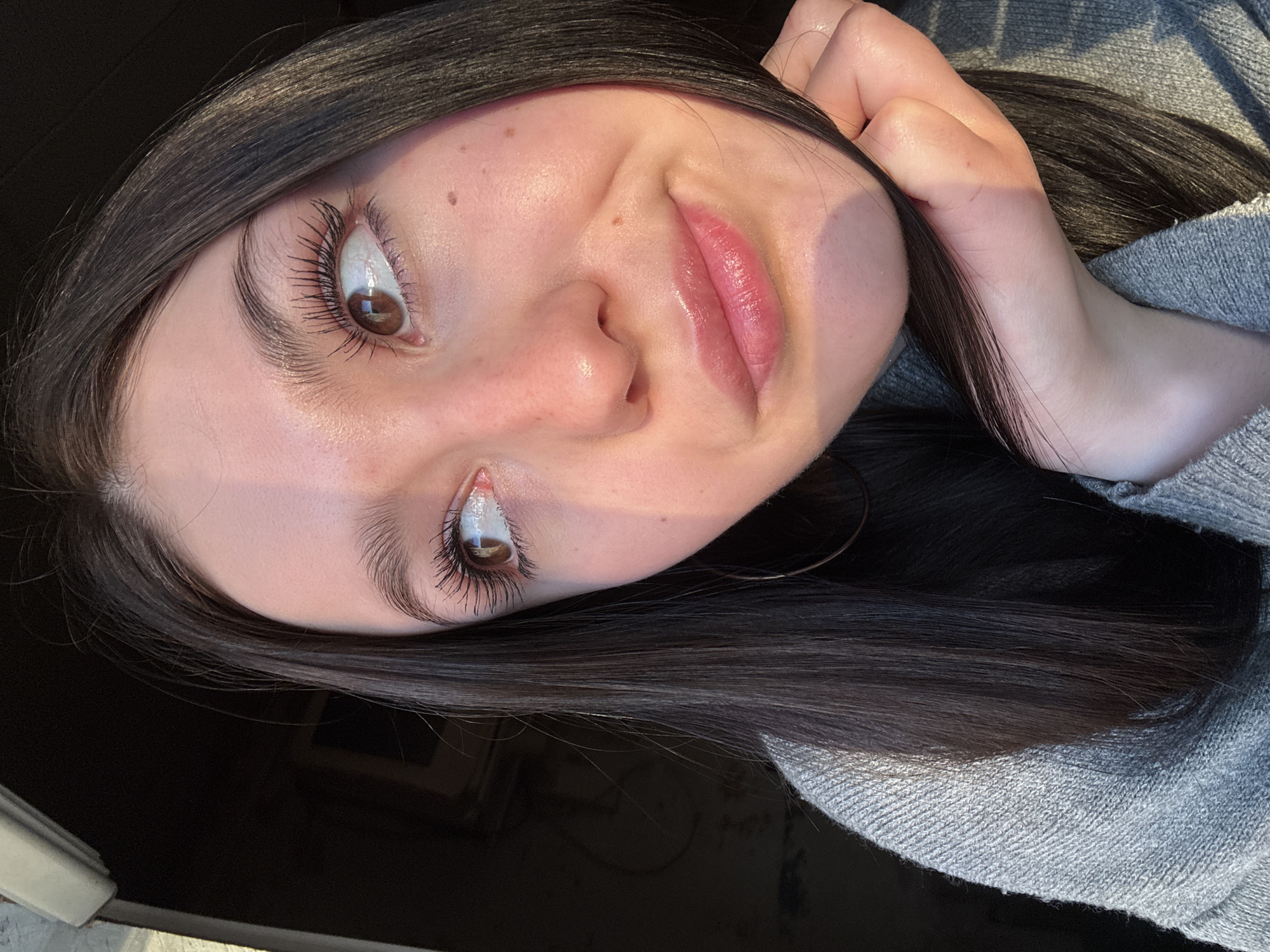 A close-up, low-angle selfie shows a young woman with fair skin and dark hair, wearing a grey sweater. Her hair is long and straight, framing her face. She is touching her hair with one hand. The woman has brown eyes with noticeable eyelashes. Her lips are glossy. The lighting suggests natural daylight. The background is a dark, undefined space. The location is not discernible.