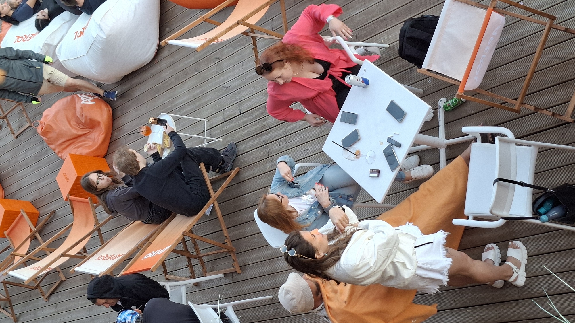 A group of people are relaxing at an outdoor cafe or lounge area with wooden decking. Several orange and white deck chairs are arranged on the deck, some occupied. There are also large orange and white beanbag chairs. A white table is set up in the foreground with several people seated around it, some with drinks and phones. A woman in a bright pink jacket is seated at the table, looking to her right. Another woman in a denim jacket is seated across from her, holding her sunglasses. A woman in a white jacket and skirt stands to the left of the table. A man in an orange shirt and a beige hat is seated near the woman in the white jacket. In the background, a man in a black hoodie and another man in a black shirt are visible, along with people seated on beanbag chairs. The overall atmosphere appears to be casual and social. The presence of "Aperol" branding on the deck chairs and beanbag chairs suggests this is an Aperol-themed establishment. The scene is consistent with an outdoor seating area in Tallinn, Estonia, likely a popular spot for leisure and socializing.