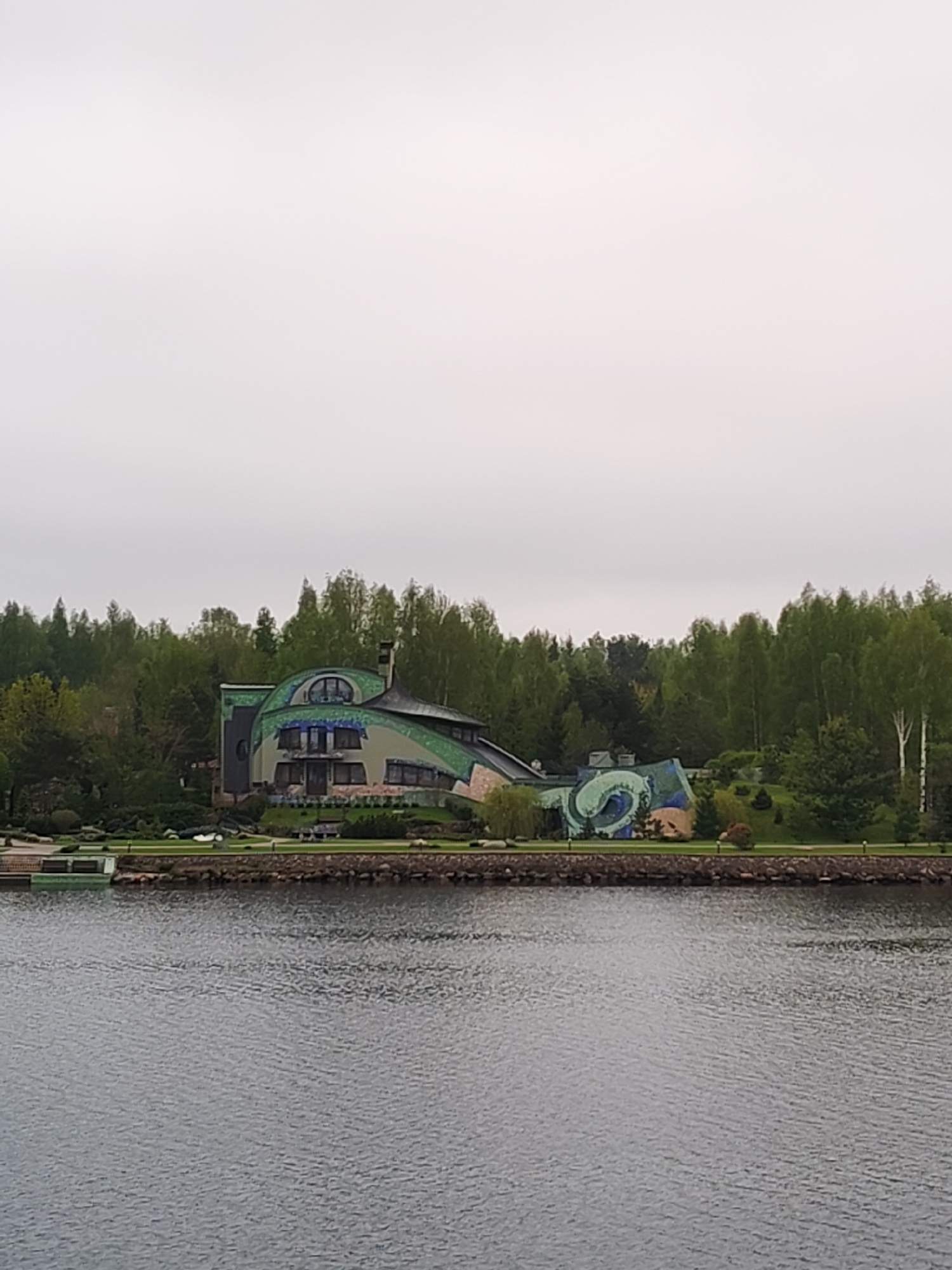 The image shows a unique, architecturally unusual house located on the shore of a body of water. The house is painted in shades of green and blue, and it features an irregular, rounded roofline and distinctive decorative elements, including a spiral design on one section. The house is surrounded by trees and landscaping. The water in the foreground is calm, and the sky is overcast.
The location of the scene is in the city of Unknown, Unknown.