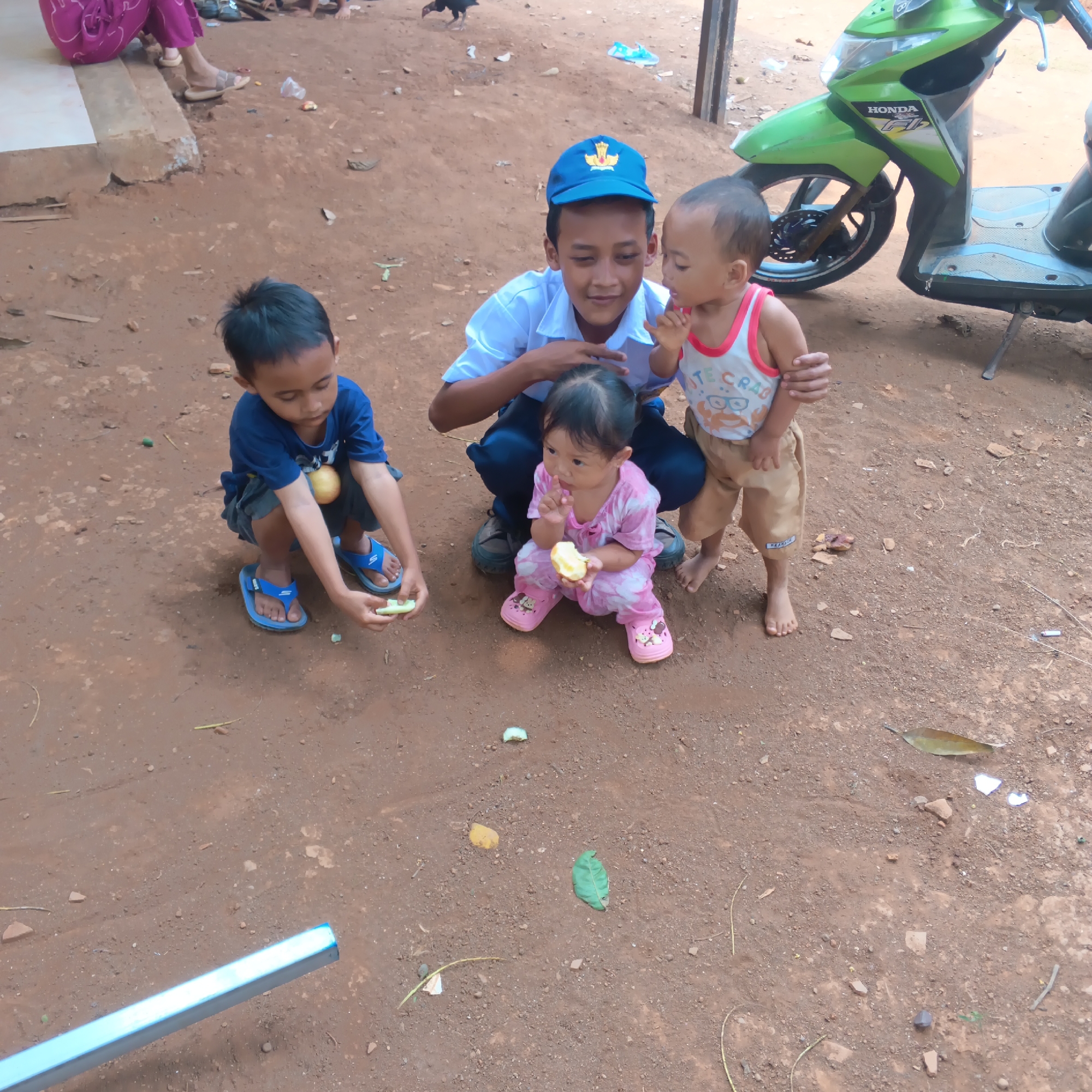 The image depicts a scene with four young children in an outdoor environment, likely in the city of Jayasampurna, Indonesia. In the foreground, two boys and a baby girl are gathered around food items on the ground. One boy, wearing a blue t-shirt and blue flip-flops, is crouching and holding a piece of fruit. Beside him, a baby girl in a pink tie-dye outfit is also holding a piece of fruit and appears to be eating. Behind them, a slightly older boy wearing a white shirt and a blue cap is kneeling, seemingly supervising. To the right, another young child in a white tank top and tan shorts stands, holding the hand of the boy in the white shirt. In the background, a green Honda scooter is parked. The ground is composed of reddish-brown dirt with scattered leaves and debris. A portion of a silver metal pipe is visible in the lower left corner of the frame. In the upper left, the lower legs of an adult wearing pink fabric are visible, seated. The overall setting suggests a casual, possibly residential, outdoor area.
