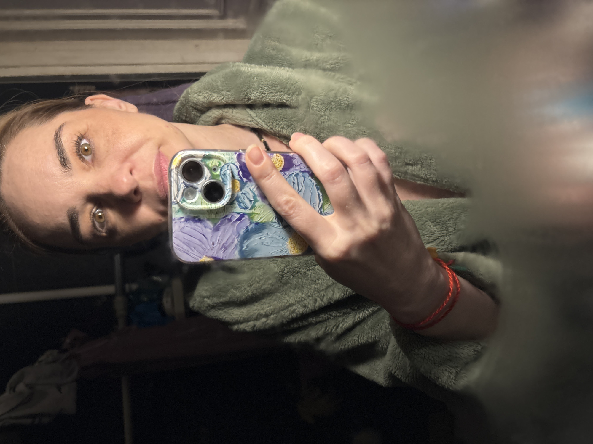 A medium close-up, slightly rotated mirror selfie shows a woman with light brown hair and prominent light brown/hazel eyes, looking directly at the camera. She appears to be in her 30s or 40s, with a subtle smile on her face, suggesting a calm or contented mood. Her eyebrows are well-defined, and she has light makeup.
She is holding a smartphone in her right hand, which covers part of her face and body. The phone has a distinct protective case featuring a colorful, floral design in shades of blue, purple, green, and yellow, resembling an impressionistic painting. The phone's three camera lenses are visible through the case's cutouts.
The woman is wearing a plush, fuzzy garment, possibly a robe or blanket, in a muted green or grey-green color. A thin, red string bracelet is visible on her left wrist. The background is dark and out of focus, indicating an indoor setting with low lighting, likely evening or night. Indistinct shapes in the background suggest furniture or other items within the room. No visible text is present in the image.