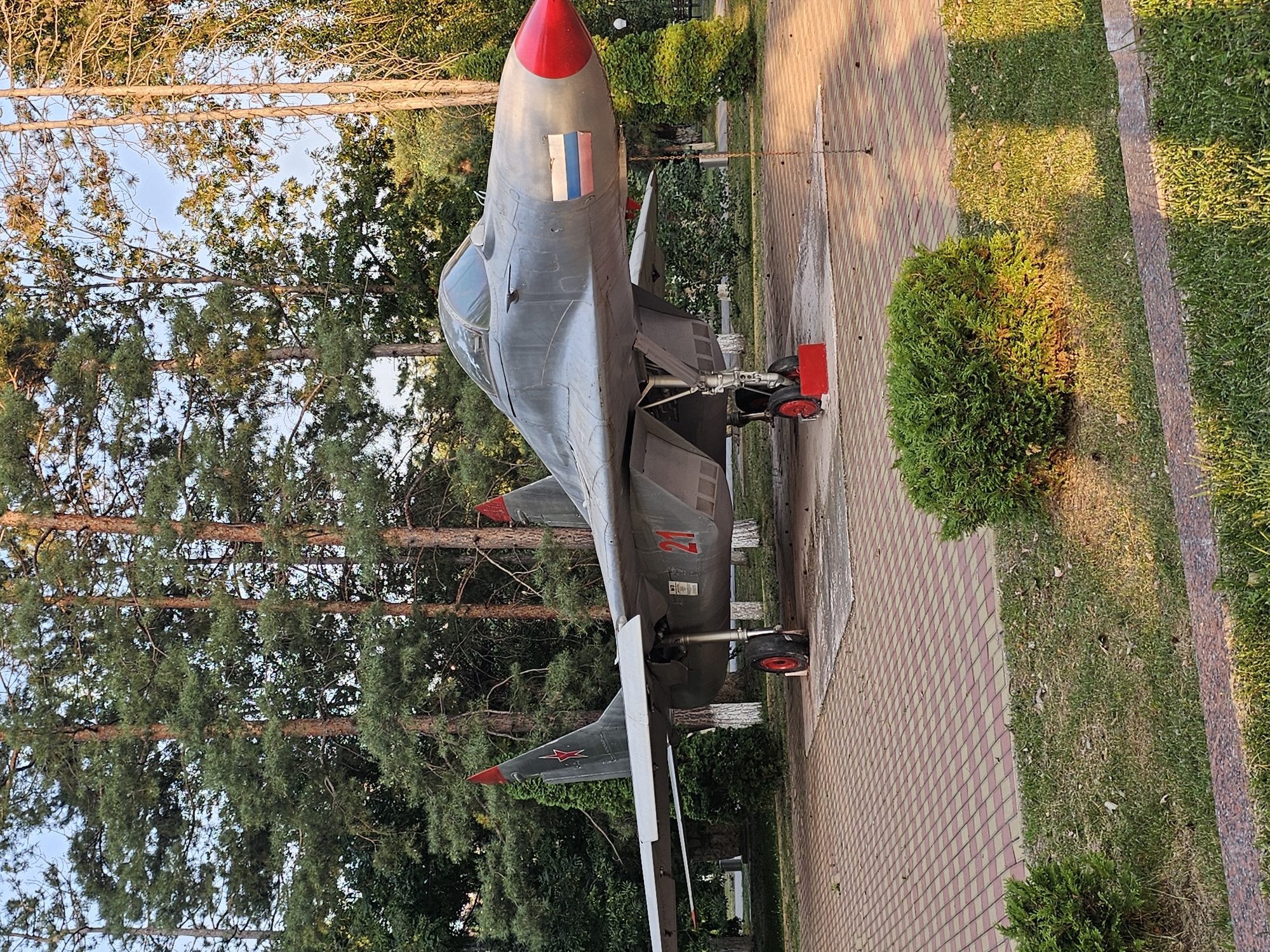 This image depicts a decommissioned Soviet-era fighter jet, identified as a MiG-29, displayed as a monument. The jet is painted in a matte gray color with red accents on the wingtips and the tip of the nose cone. A Russian flag decal is affixed to the fuselage near the nose. The number "21" is prominently displayed in red on the side of the aircraft. The jet is positioned on a paved area made of interlocking red and yellow tiles, which is surrounded by a patch of green grass and manicured bushes. Tall pine trees form a dense backdrop to the scene. The setting appears to be an outdoor park or public space. Based on available information, this monument is located in the city of Cherkessk, Russia. There are no people or animals visible in the image. The lighting suggests it is daytime, with sunlight casting shadows.