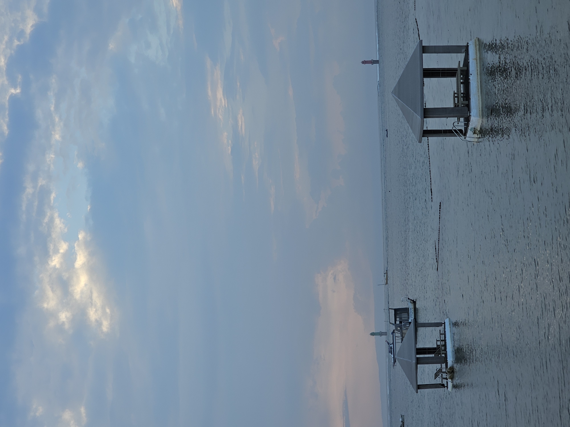This is a vertically oriented, eye-level shot of a calm sea at dusk, possibly Sanur Beach in Denpasar, Indonesia, due to the presence of floating gazebos and lighthouses often seen in that area. The sky is filled with muted blue and grey clouds, with hints of pink and yellow near the horizon, indicating the sun is setting or has just set. Two hexagonal floating gazebos are prominent in the foreground and middle ground, each with a simple grey roof supported by pillars, a small deck, benches, and a table inside. The gazebo on the right appears to have a ladder for entry. The water is a deep grey-blue with gentle ripples and reflections of the gazebos. In the distance, there are several boats, including a larger boat with the name "SANTRAN" visible, a tall green lighthouse, and a red lighthouse on the horizon, suggesting a navigational area or harbor entrance. The overall atmosphere is serene and peaceful, with a low level of activity. No people are visible in the frame.