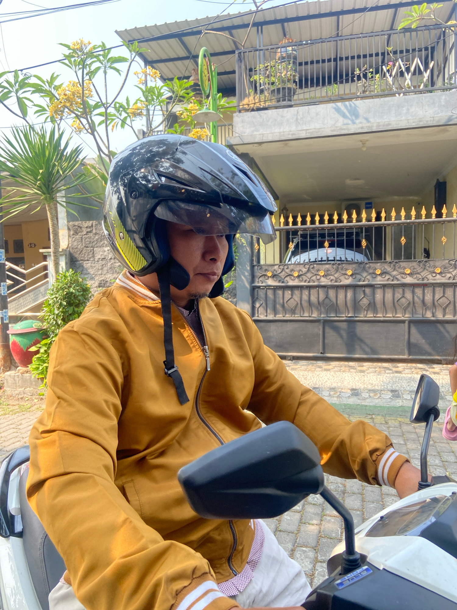 A medium shot captures a man wearing a black helmet and a tan jacket while sitting on a white motorcycle. The motorcycle is parked on a paved surface in front of a house with a fence. The man appears to be looking forward, with his hands on the handlebars. A palm tree and a flowering bush are visible to the left of the motorcycle. The background shows a two-story house with a balcony and a metal fence. The scene is illuminated by daylight. The location is identified as Gelam, Indonesia.