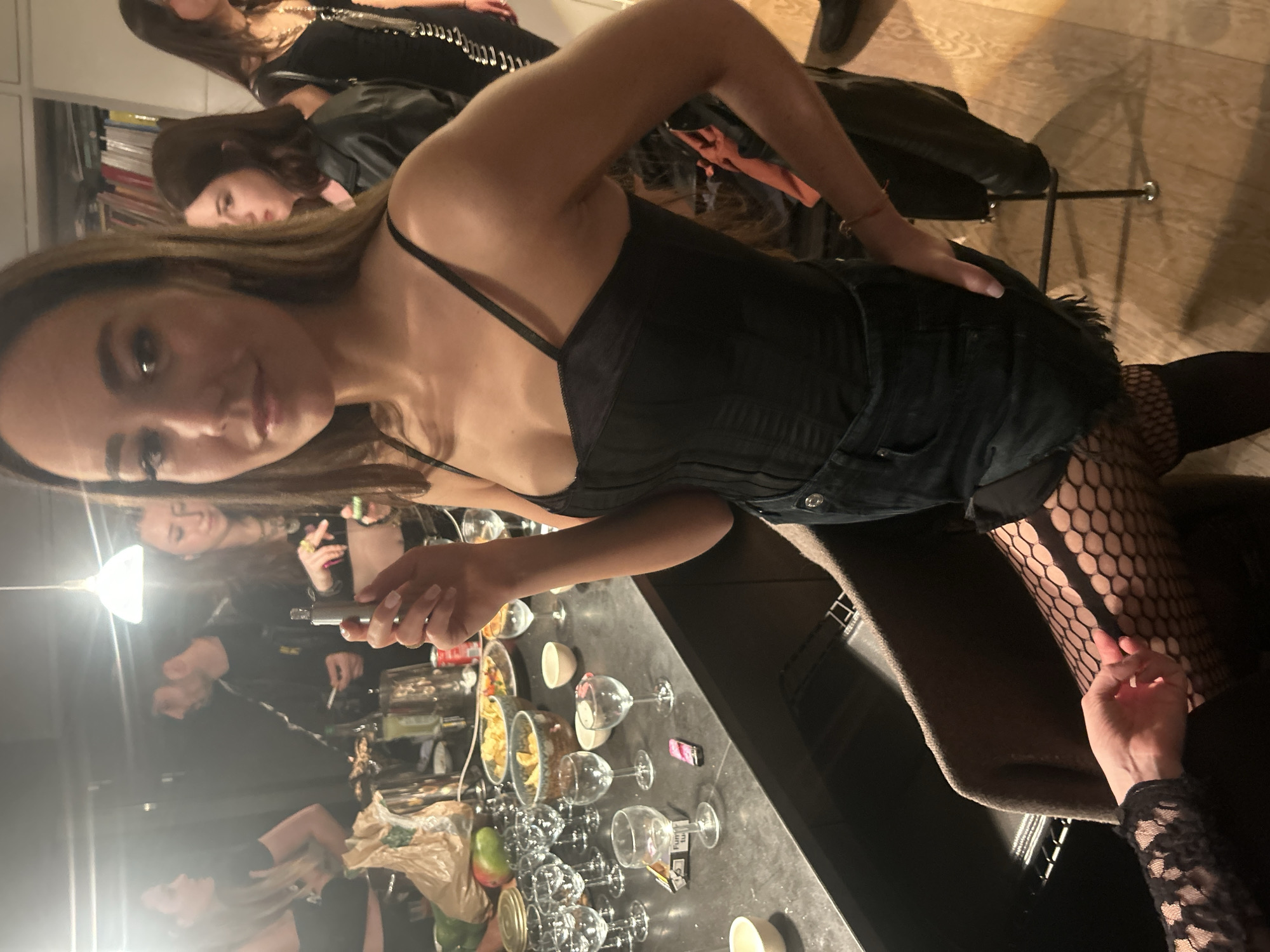 The image depicts a social gathering of multiple individuals in what appears to be an indoor setting, possibly a private residence or a venue with a bar area. The main subject, a woman with brown hair, is in the foreground wearing a black corset top, denim shorts, and fishnet stockings. She is holding a small object in her hand, possibly a cigarette or vaping device. To her right, a hand wearing a black lace sleeve is visible.
The scene includes a bar counter or table laden with several wine glasses, bowls of chips and snacks, and other items, suggesting a casual party environment. Multiple people are in the background, some holding what appear to be cigarettes, indicating smoking or vaping is occurring within the gathering. There is some clutter visible, including a shopping bag. Other individuals wear dark clothing, including leather jackets. A bookcase is visible in the back. The specific location in the city of Unknown, Unknown is unidentifiable based on this image.