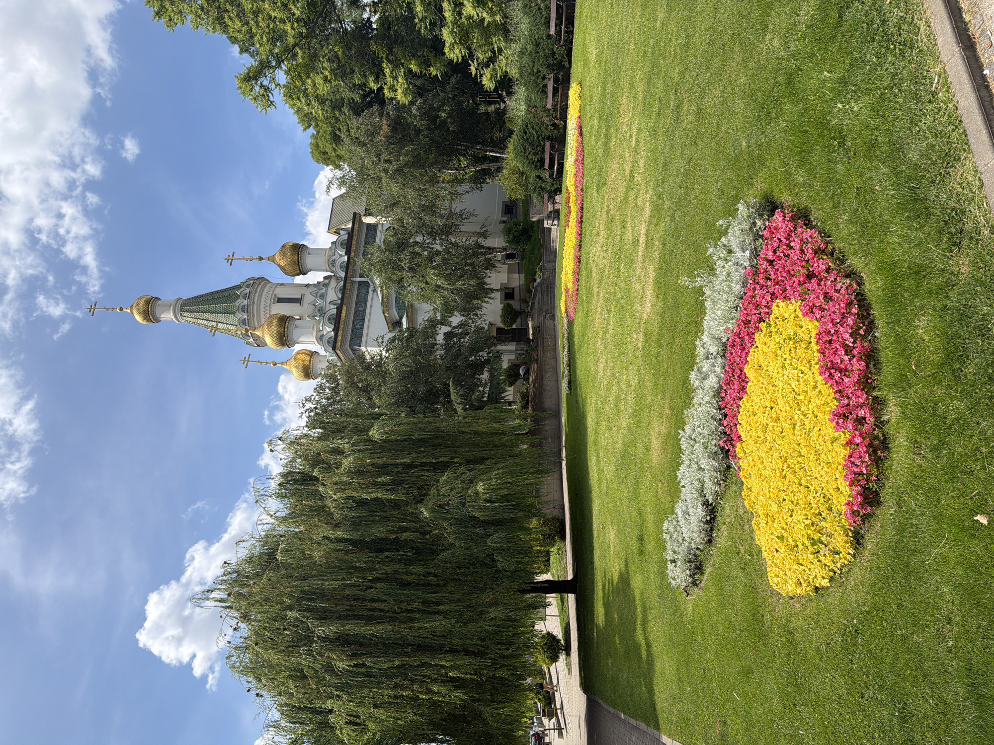 The image depicts the St. Nicholas the Miracle-Maker Russian Church, a landmark located in Sofia, Bulgaria. The church, characterized by its distinctive green and gold domes, stands prominently against a bright blue sky dotted with white clouds. In the foreground, a well-maintained lawn is adorned with vibrant flower beds in circular patterns, featuring yellow, pink, and silver foliage. A large weeping willow tree with cascading branches stands to the left of the frame, casting a shadow on the grass. Several dark wooden benches are visible in the middle ground, facing the flower gardens. The scene is set within a park-like environment, with paved walkways, other trees, and bushes. The overall atmosphere is serene and picturesque, capturing a sunny day in the city.