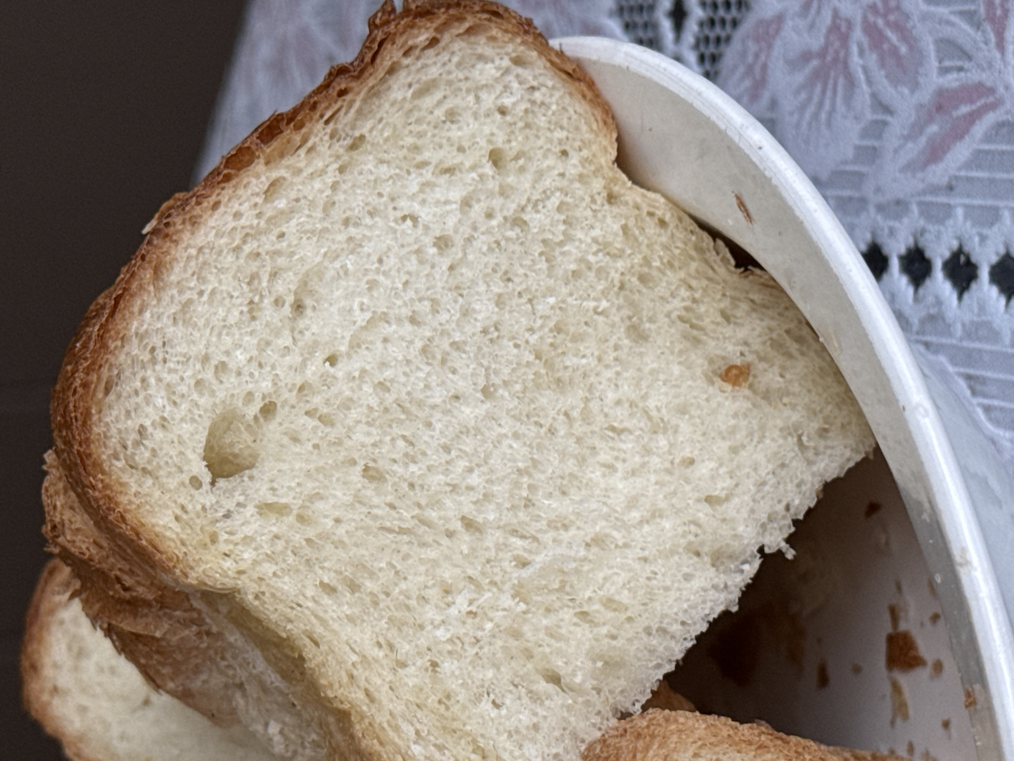 A close-up photograph shows a stack of sliced white bread in a white plastic container. The crust is golden brown. The background consists of a table covered with a white lace tablecloth. The image provides no information about the location in Верхнеказымский, Russia, or any people, subjects, objects, actions, interactions, or environment beyond what is visible in the frame.