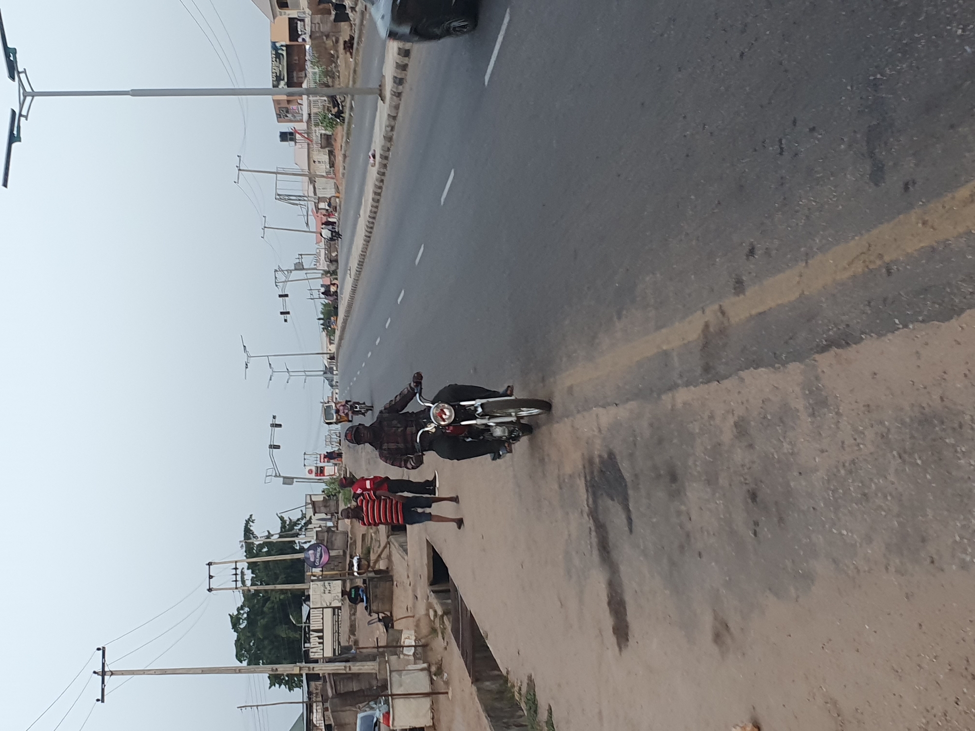 The image depicts a street scene in Lokoja, Nigeria. A motorcyclist is traveling on a paved road with painted lane markings. Two pedestrians are walking along a concrete sidewalk next to the road. Utility poles and streetlights line the side of the road. Buildings and trees are visible in the background. The sky is overcast.