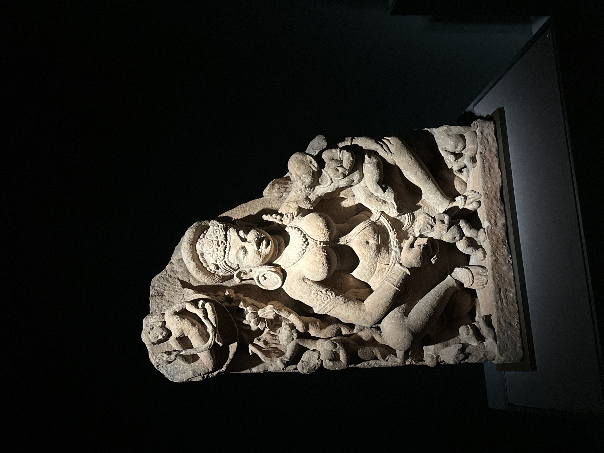 This is a medium shot, eye-level photograph of a stone sculpture of a woman with multiple figures surrounding her. The sculpture is displayed on a dark gray pedestal in a dimly lit room. The background is black, creating a dramatic contrast with the light that illuminates the sculpture. The sculpture depicts a central female figure with prominent breasts and elaborate jewelry, including a headdress, earrings, and necklaces. She is in a kneeling or squatting position. Around her are smaller figures, some of which appear to be children, clinging to her or interacting with her. The sculpture is carved from a light-colored stone, possibly sandstone, and shows signs of age and wear. The lighting is directional, highlighting the details of the carving and casting shadows that add depth and drama to the scene. There is no indication of the specific location in Seoul, South Korea, in the image itself.