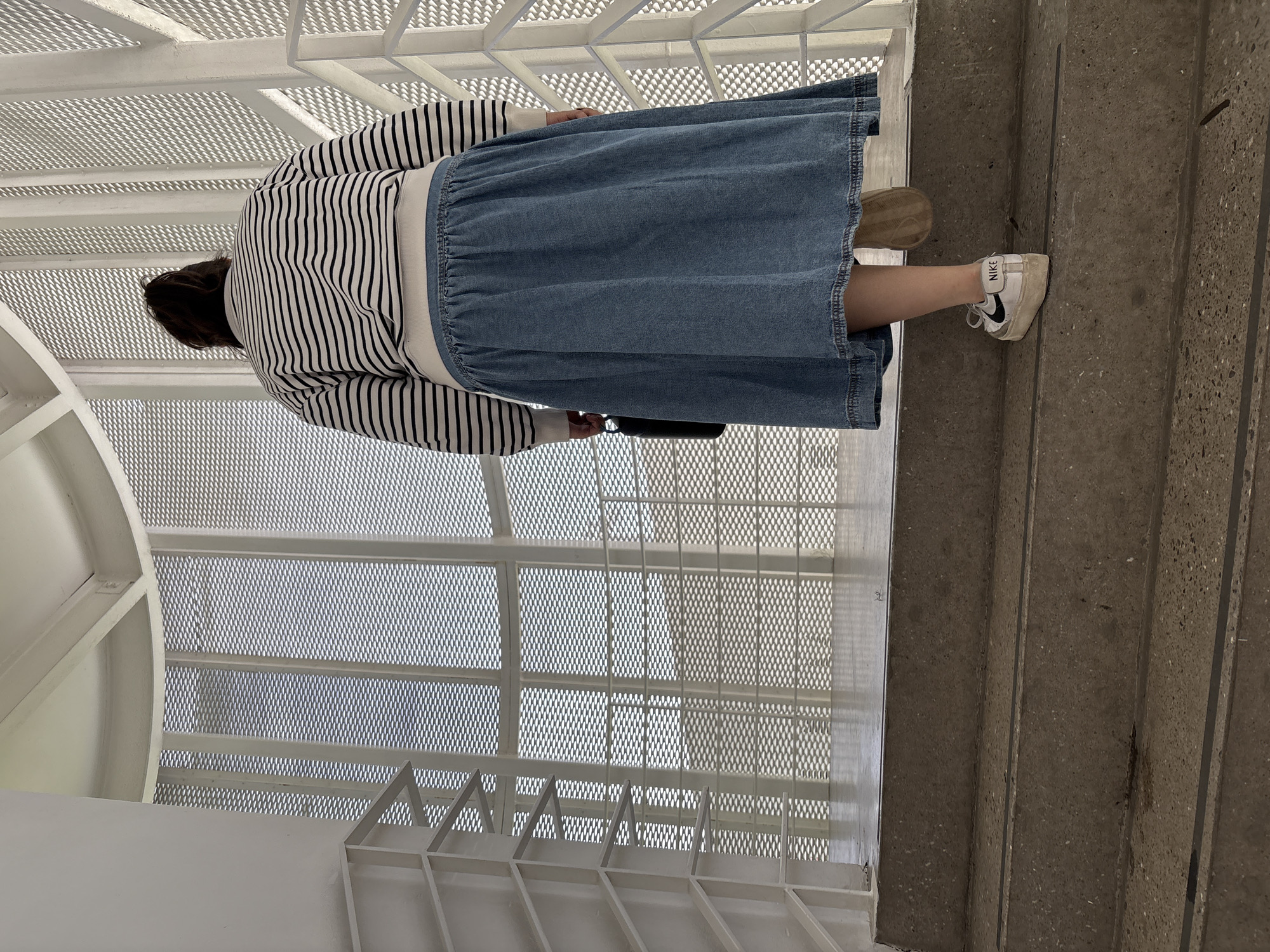 A high-angle shot shows a person walking up concrete steps beside a metal staircase. The person is wearing a striped long-sleeved shirt, a denim skirt, and white Nike sneakers. They are holding a black item, possibly a bag. The setting includes a white metal structure with a mesh-like design visible through it, likely part of the staircase or surrounding architecture. The steps are a light gray concrete and have straight lines.
