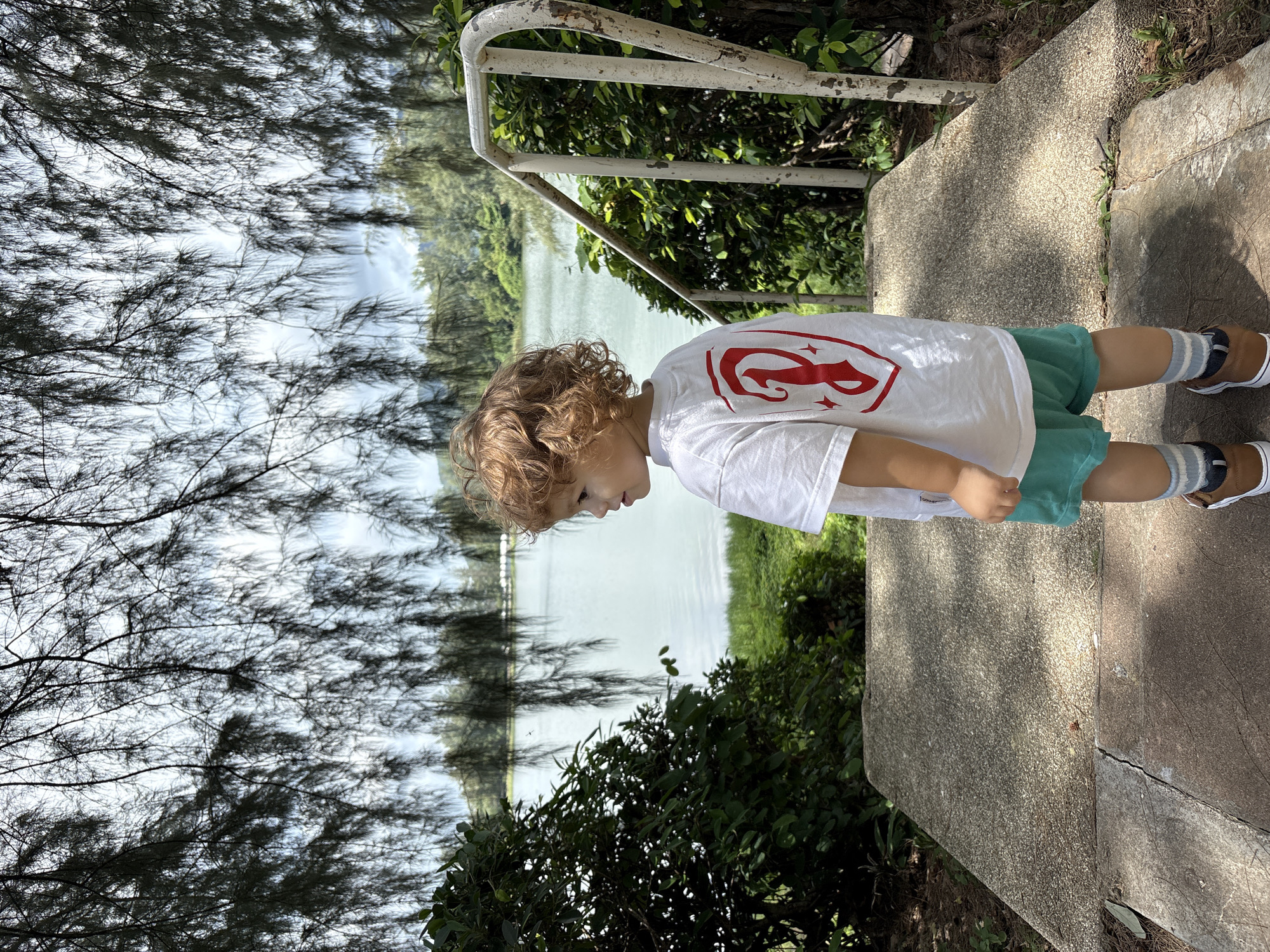In Choeng Thale, Thailand, a young child with curly blonde hair stands facing away from the camera, looking towards a body of water. The child is wearing a white t-shirt with a red graphic on the back, turquoise shorts, striped socks, and brown shoes. They are positioned on a concrete pathway that leads to a set of outdoor stairs with a white railing. The pathway and stairs are partially shaded by the branches of trees overhead, with lush green foliage surrounding the area. The background features a calm, pale blue lake or pond with trees and vegetation on the opposite shore. The sky is overcast with some patches of blue visible.