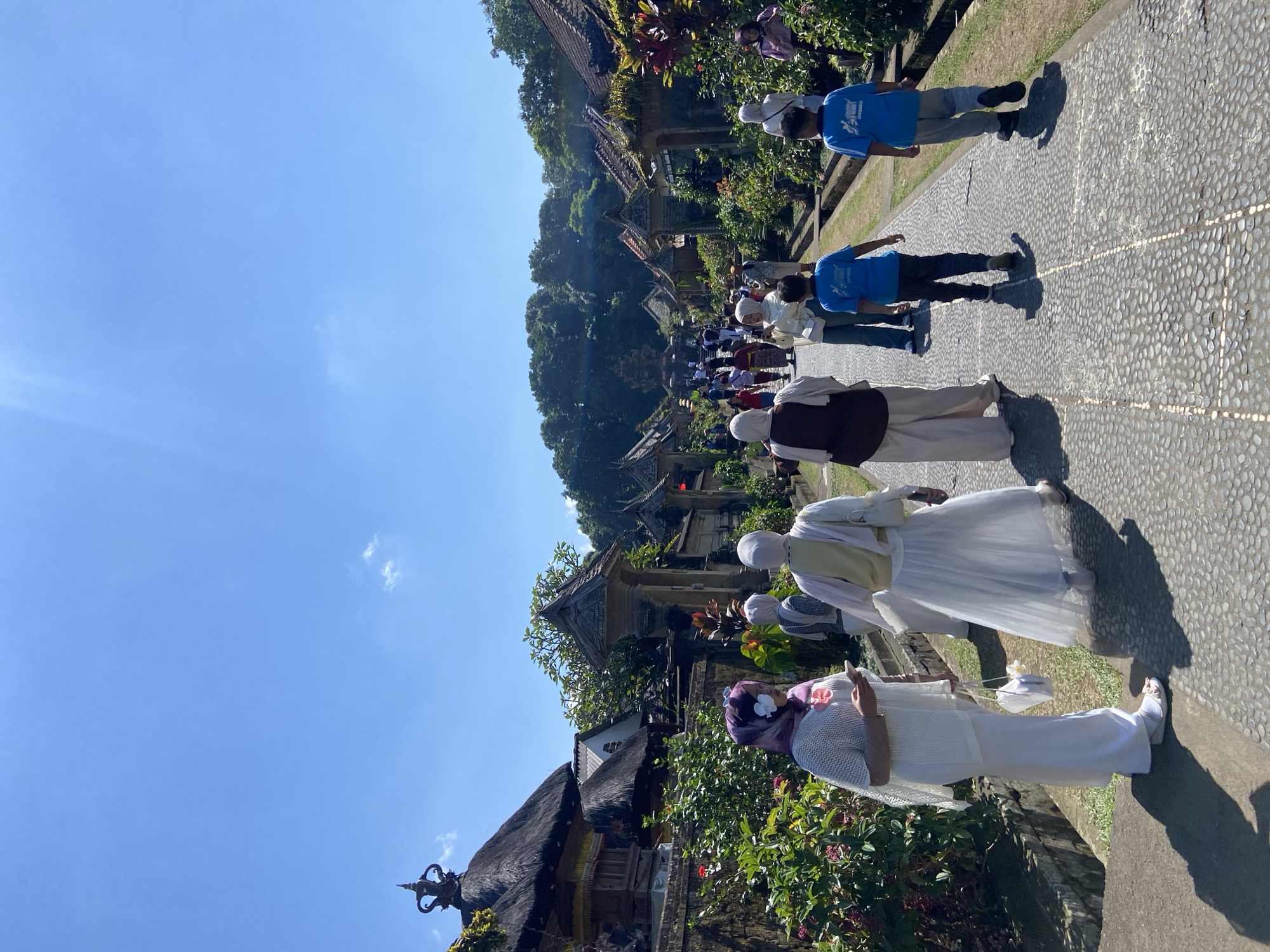 The image depicts a sunny day in Bangli, Indonesia, with a clear blue sky overhead. A group of people, many dressed in light-colored, traditional clothing including hijabs and long skirts, are walking along a cobblestone path. The path is lined with lush greenery, flowering plants, and traditional Balinese architecture with thatched roofs. In the background, more people are visible walking towards what appears to be a ceremonial structure, suggesting a temple or cultural site. The overall scene conveys a sense of community and cultural engagement in a scenic, historic setting.