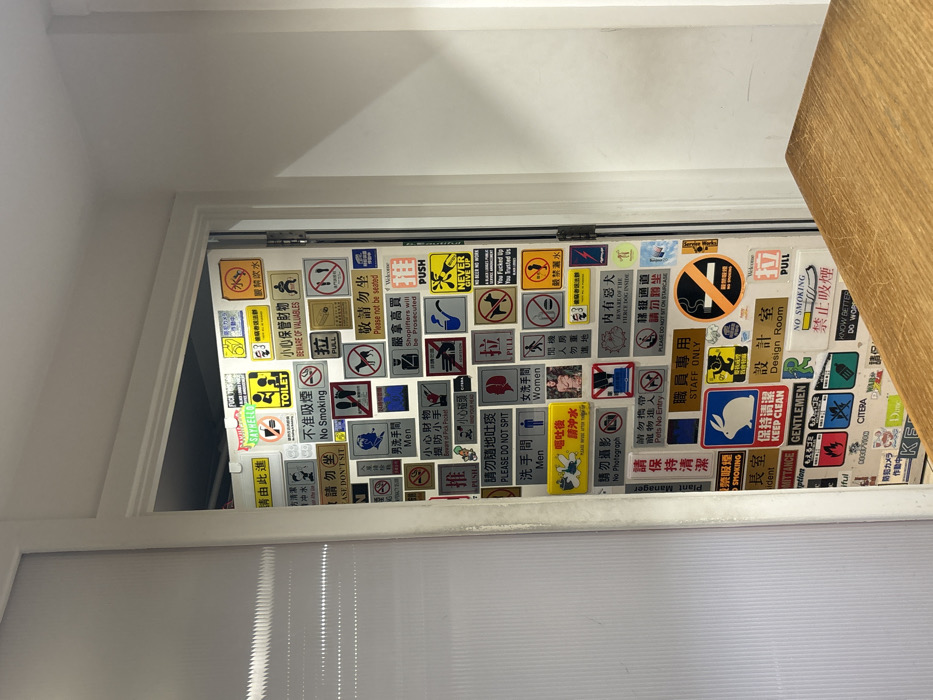 The image displays an interior view of a white door almost entirely obscured by a dense collage of approximately one hundred assorted signs. These signs vary in shape (rectangular, square, circular), color, and font, featuring text predominantly in traditional Chinese characters and English. The content encompasses a diverse range of messages, including prohibitions such as "No Smoking," "Please Do Not Spit," "No Photograph," and "Pets No Entry," alongside warnings like "Beware of Valuables," "Beware of Pick Pocket," and "Shoplifters will be prosecuted." Instructional signs include "Pull," "Push," "Toilet," "Men," and "Women." Interspersed are informal or humorous declarations such as "FUCK OFF," "SAY HELLO," and "You Fucked Up You Trusted Us." Icons depict figures, animals (e.g., a rabbit, a dog), and common prohibition symbols. To the immediate left of the door frame, a vertical, translucent, ribbed white panel is visible. A plain white wall is seen to the right, and a light brown wooden tabletop occupies the lower right quadrant of the foreground. This specific configuration of numerous, eclectic bilingual signs is characteristic of the Misato restaurant located in Leicester Square, London, United Kingdom.