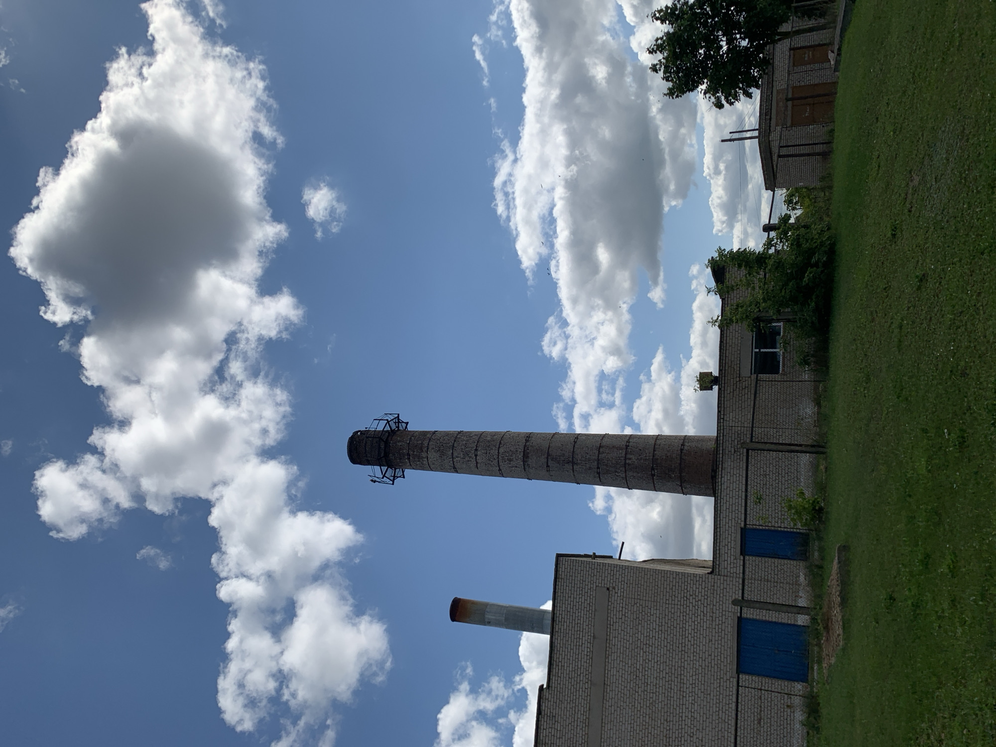 The image depicts an industrial or factory setting against a bright blue sky with scattered white clouds. Two prominent brick chimneys rise from the complex, the taller one being a cylindrical structure with a metal platform near its top. To the left of the taller chimney, a smaller, narrower metal chimney with a rusted top extends from a brick building. This building, constructed with light-colored bricks, features two blue doors and is enclosed by a wire fence. Adjacent to this building, another smaller brick structure with brown doors is visible, partially obscured by greenery. The ground in the foreground is covered with green grass. The scene is devoid of any people or animals. The location is identified as the city of Kapyĺ, Belarus.