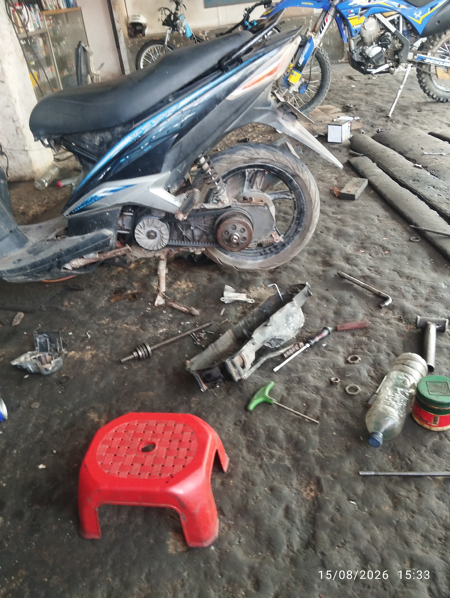 This is an outdoor, low-angle, medium shot of a motorcycle repair scene. The environment is a concrete floor littered with debris, tools, and motorcycle parts, suggesting a workshop or an outdoor repair area. In the foreground, a bright red plastic stool is positioned on the ground. To the left of the stool, a black and blue scooter is partially visible, with its rear wheel and engine exposed. Further back and to the right, a blue and white dirt bike is parked. Scattered around the scene are various tools, including wrenches, a screwdriver, and a green T-handle tool. A plastic water bottle and a green tin are also present on the ground. The lighting suggests it is daytime. The timestamp in the lower right corner indicates the image was taken on August 15, 2026, at 15:33.