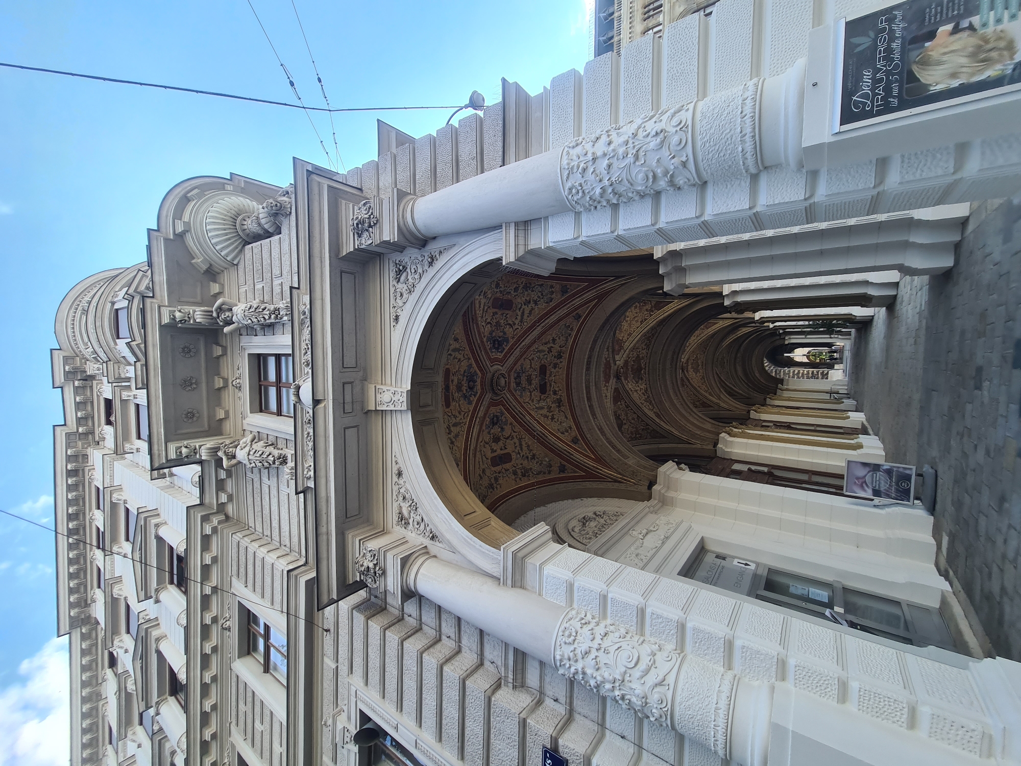 The image shows a low-angle, wide shot of a grand arched walkway leading into a building on a sunny day in Vienna, Austria. The building's facade is ornate, with white stone detailing, intricate carvings, and multiple windows. The walkway itself is covered by a vaulted ceiling adorned with colorful decorative patterns. White columns with floral carvings line the sides of the walkway.
On the left side of the walkway, a glass door is visible with signage above it that reads "SALONSA" and "ENGANG" which means "Entrance" in German. A poster is propped up outside this entrance.
On the right side of the walkway, near the foreground, a sign is displayed with text that appears to be an advertisement for a hair salon. The sign includes an image of a woman with blonde hair and text in German, "Deine Traumfrisur ist nur 5 Schritte entfernt" which translates to "Your dream haircut is only 5 steps away."
The walkway leads into a dimly lit interior with more arches visible in the distance. The ground of the walkway is paved with grey cobblestones. Overhead, blue sky with some clouds is visible, along with power lines. The overall impression is one of architectural grandeur and a bustling urban environment.