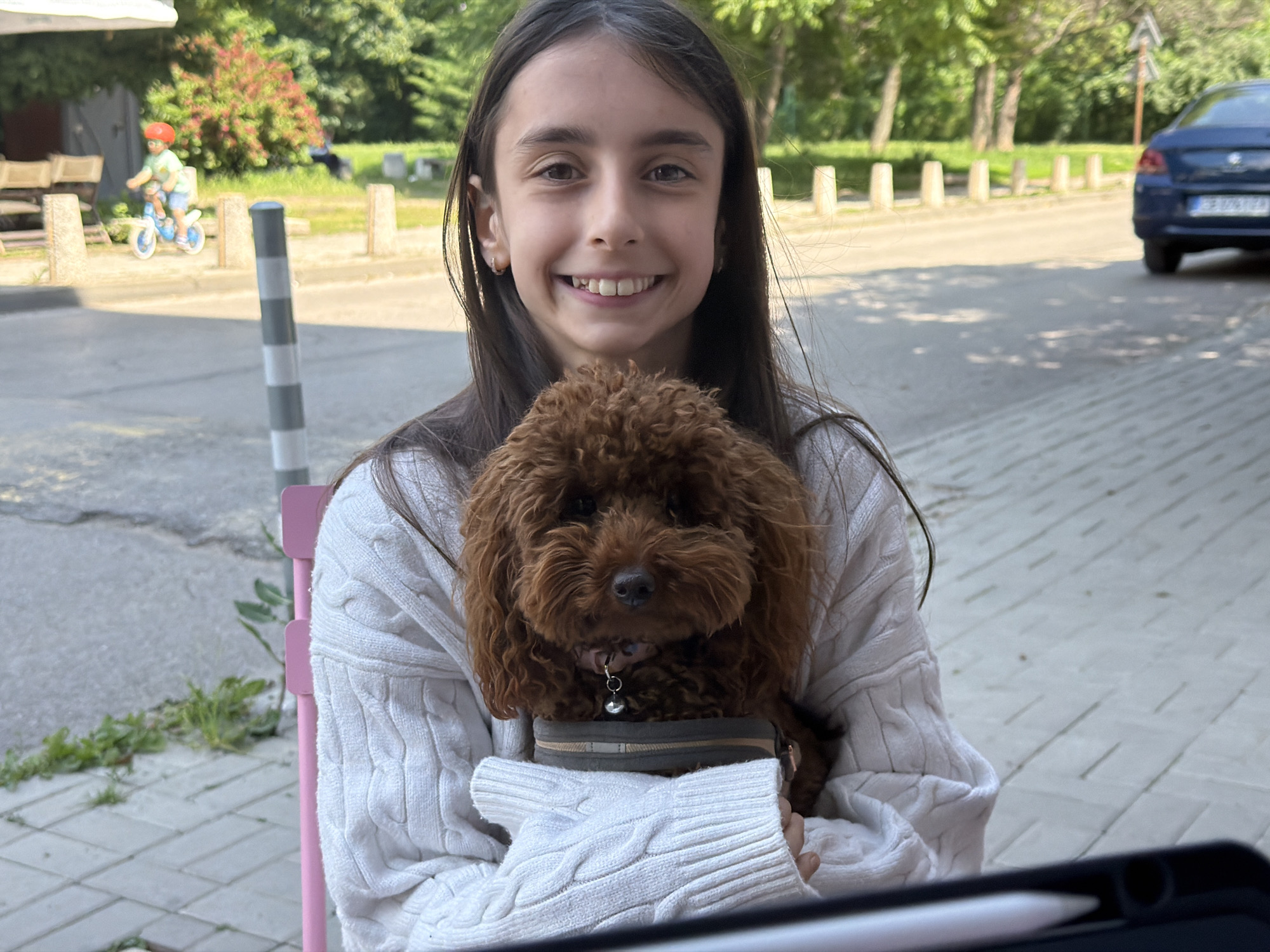 The image displays an outdoor scene under bright daylight, located in Sofia, Bulgaria, identifiable by the "CB" region code visible on a vehicle's license plate. In the foreground, a young female with long dark hair and a wide, open-mouthed smile, exposing her teeth, is seated, facing the viewer directly. She is wearing a white cable-knit sweater. Cradled in her arms or lap is a small, curly-haired, reddish-brown dog, possibly a poodle, which is also looking towards the viewer. The dog wears a collar with a small silver bell attached and appears to be positioned within a grey and tan carrying device. A portion of a pink chair is visible behind the girl to her left. In the lower right, a black tablet case and a white stylus are partially visible on an unseen surface.
The midground features a paved area, likely a sidewalk or plaza, bordered by a series of light-colored, short bollards. Beyond these bollards lies an asphalt road. To the left of the central subjects, a young child wearing an orange helmet and light-colored attire is seen riding a small blue balance bike. Further left, elements of an outdoor cafe are discernible, including a white market umbrella and wooden tables and chairs. To the right, a blue sedan is parked or in motion on the road, with its license plate showing "CB" followed by digits, confirming the Bulgarian location. The background throughout the scene is dominated by lush green trees and bushes, indicating a park-like or heavily treed environment. Shadows are cast across the paved surfaces, consistent with a sunny day.