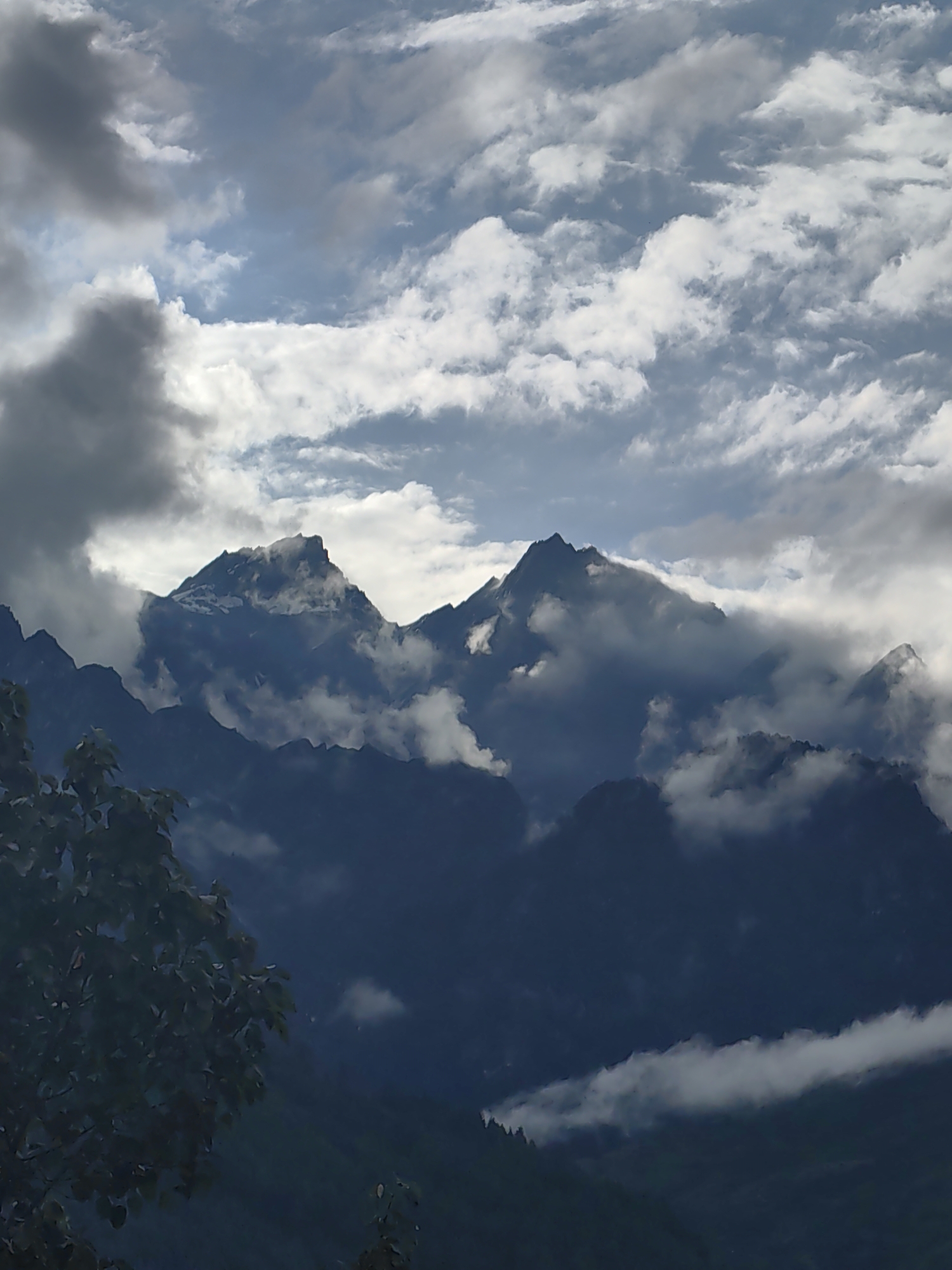 A photograph captures a majestic mountain range in Vashisht, India, under a dramatic sky. The peaks are shrouded in swirling clouds, some dark and ominous, others illuminated by the sun. Snow-capped summits peek through the mist, hinting at the immense scale of the mountains. In the foreground, the dark silhouette of foliage frames the scene, adding depth and contrast. The lighting suggests either early morning or late afternoon, with the sun casting a diffused glow through the clouds. The overall impression is one of raw, natural beauty and the awe-inspiring power of the elements.