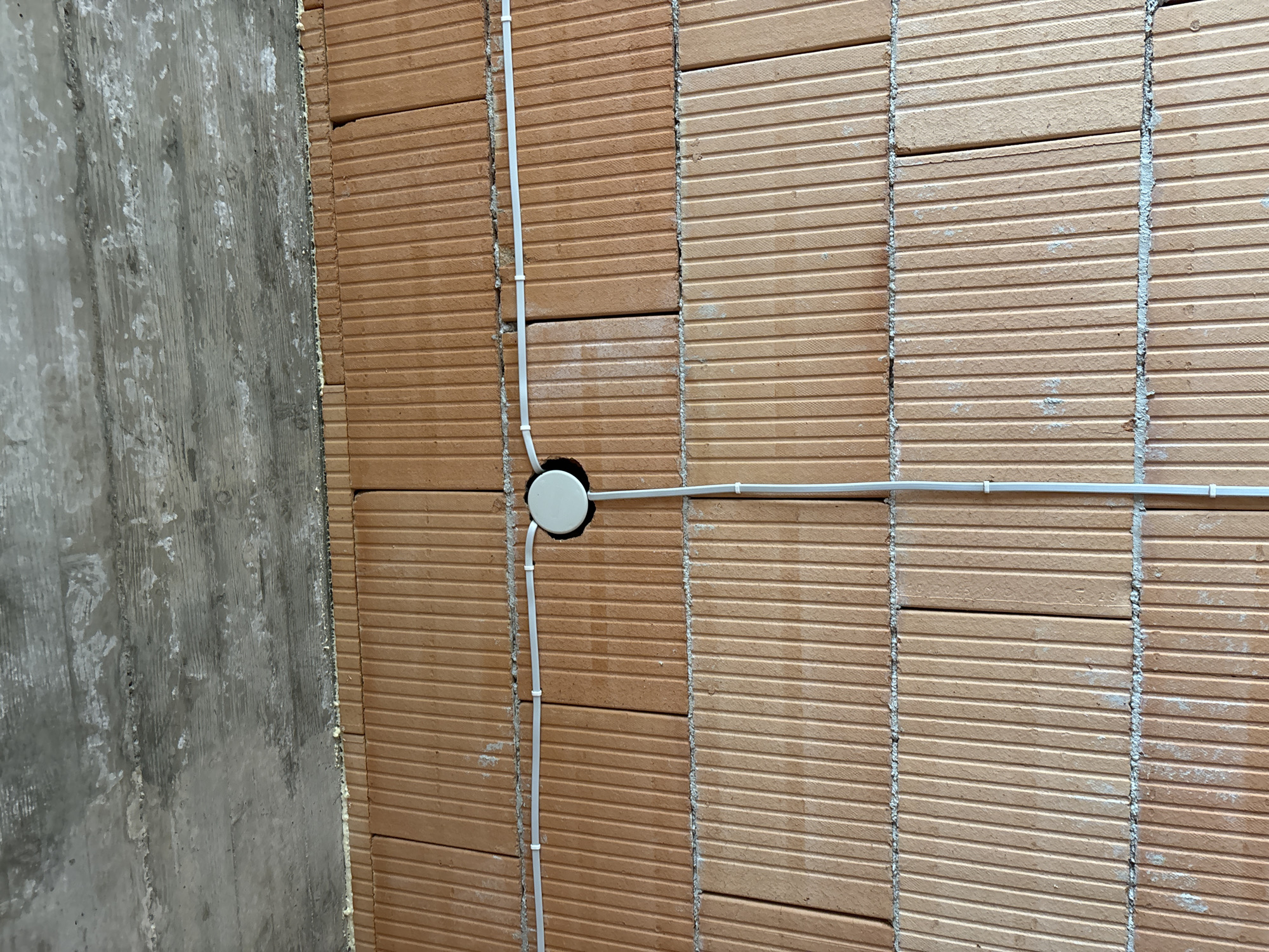 The image depicts an interior wall under construction. The wall is made of reddish-brown clay blocks with horizontal grooves. The blocks are laid in rows, with mortar visible in the vertical joints between the blocks. A section of a gray concrete wall is visible on the left side of the image. White electrical conduits are attached to the clay block wall. One conduit runs vertically, bends, and enters a white circular junction box mounted on the wall. Another conduit exits the junction box and runs horizontally along the wall. The electrical conduits are secured to the wall with plastic clips. There is no evidence of people, actions, or interactions within the scope of this image. There are no explicit indicators of the city of Burgas, Bulgaria.