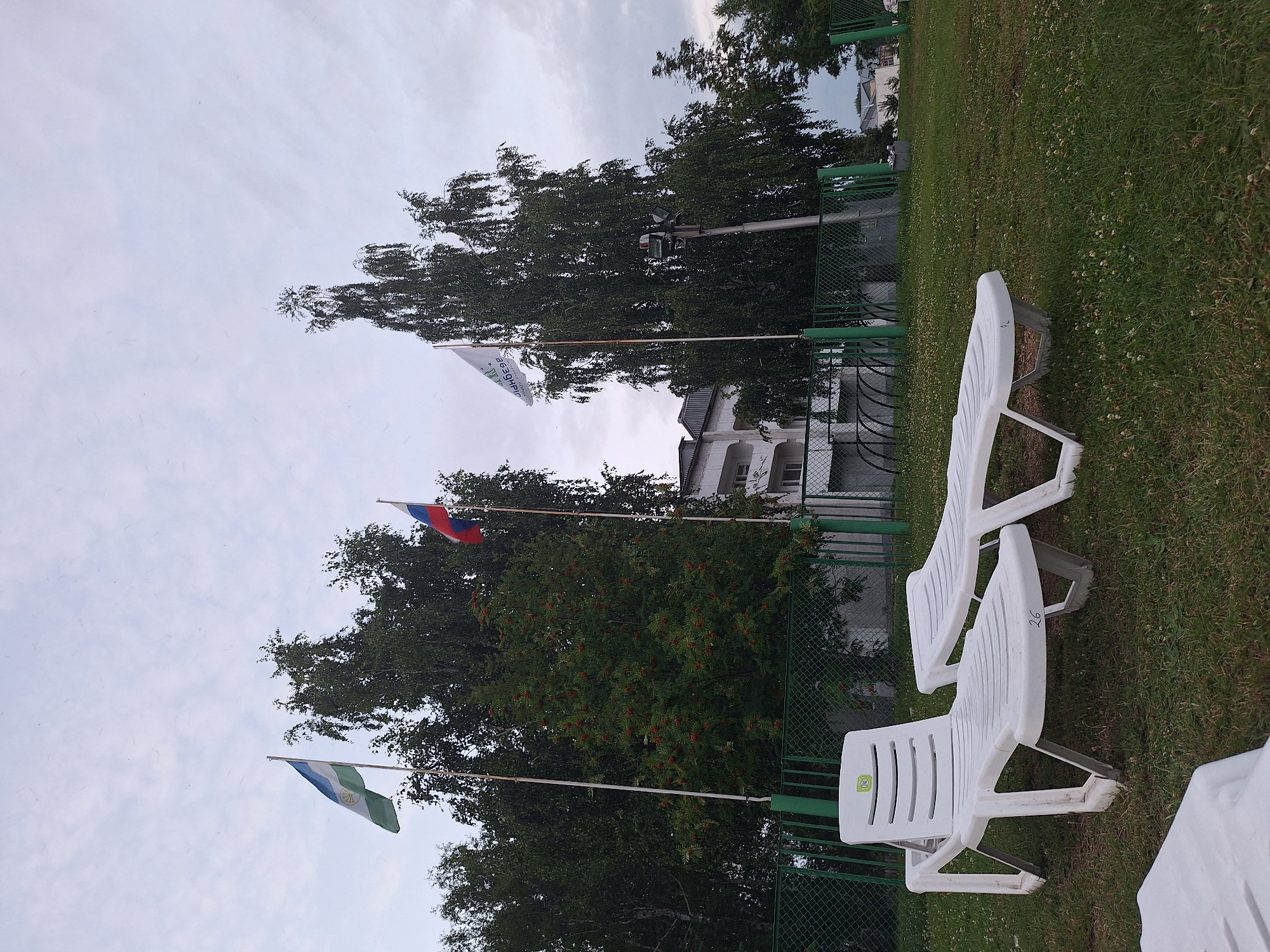 The image depicts an outdoor scene on an overcast day. In the foreground, two white plastic lounge chairs are positioned on a grassy lawn. One chair, numbered "26", is closer to the viewer and angled slightly. Behind it, another lounge chair is partially visible. A third white object, possibly another lounge chair, is seen in the bottom left corner. A green chain-link fence runs horizontally across the middle ground. Behind the fence, several trees with dense foliage are visible, one of which is laden with red berries. Three flags are flying on poles in front of the trees. The leftmost flag is tricolored with blue, white, and green. To its right, a flag with red and white stripes and a blue stripe is visible. Further to the right, a white flag with text and a logo is displayed. In the background, a multi-story building with balconies and windows can be seen, along with more trees and a distant structure. The overall environment suggests a public outdoor recreational area, possibly within the city of Atamanovka, Russia. There are no people or animals present in the image.