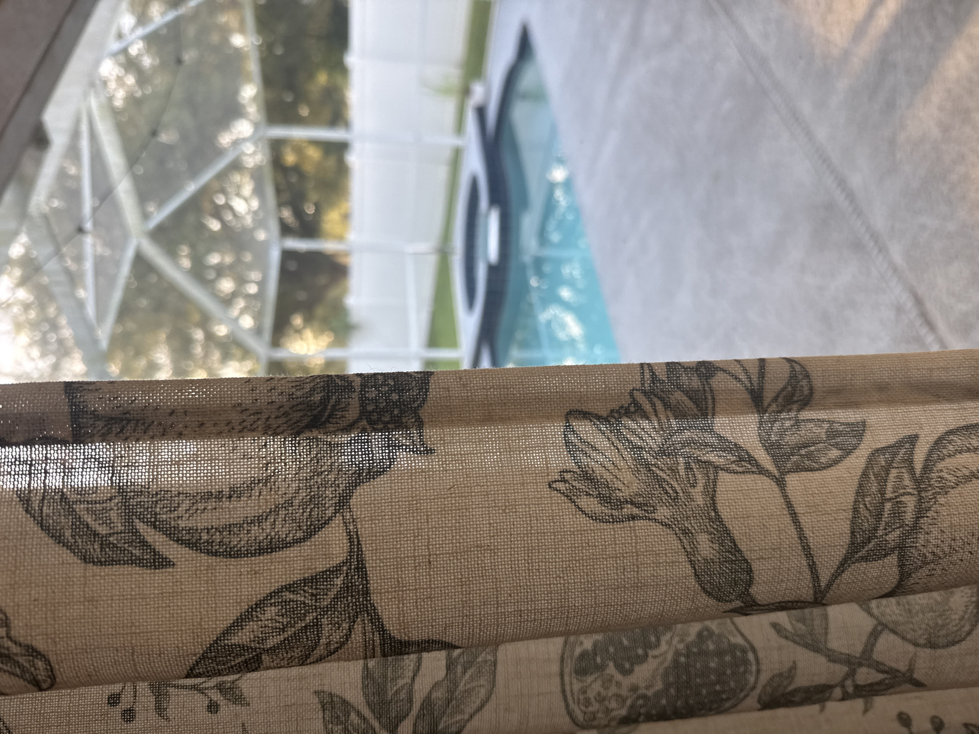 The image shows a close-up of a patterned curtain in the foreground, with a screened-in swimming pool and spa in the background. The curtain has a botanical print featuring leaves and what appear to be pomegranates. Sunlight is filtering through the curtain and the screened enclosure, creating a dappled light effect. The swimming pool is a bright turquoise color, and the spa appears to be integrated into the pool area. A white fence surrounds the outdoor space. The scene is likely a residential backyard in Trinity, United States of America.