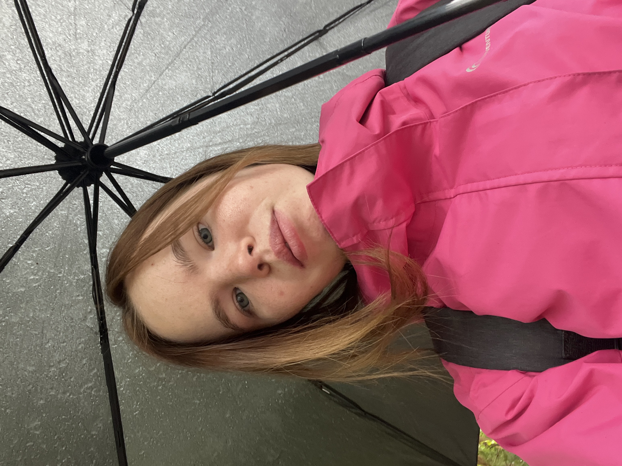A young woman is under an open umbrella. She is wearing a pink jacket with a black strap, possibly a backpack strap, visible over her shoulder. The umbrella is dark gray, covered in raindrops. The spokes of the umbrella are black. Her hair is light brown, with sun-kissed highlights. Her eyes are blue. She appears to be looking at the camera. The location is not explicitly identifiable as Chaikovsky, Russia, based on the visual information in the image.