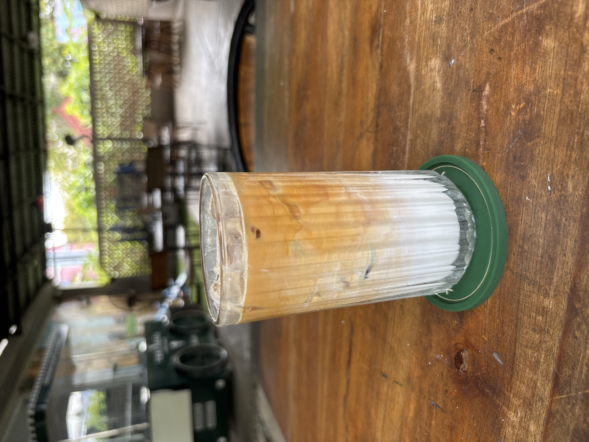 A glass of iced coffee sits on a wooden table, a green coaster underneath. The coffee is layered, with a light cream base topped with darker coffee. The table appears to be outdoors, given the blurred background of greenery and building structures. The image was taken in Kuta Selatan, Indonesia. There are no people visible in the frame.