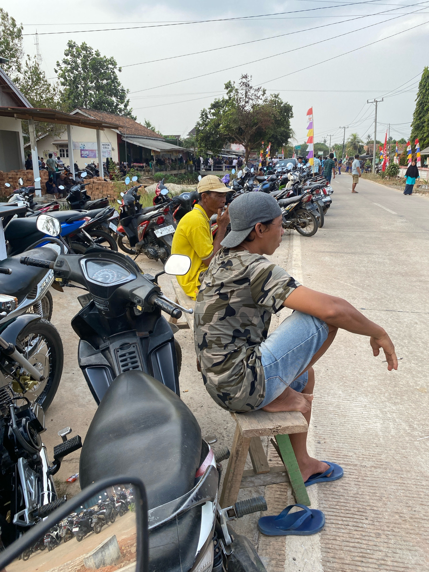 The image depicts a street scene in Sri Sawahan, Indonesia, on an overcast day. In the foreground, a man wearing a camouflage t-shirt and denim shorts is seated on a wooden stool, holding a cigarette. Next to him, another man in a yellow t-shirt is also seated, facing away from the camera. A row of parked motorcycles lines the street, extending into the background. Buildings with corrugated roofs and awnings are visible on the left side of the street, with people gathered in front of them. On the right side, more motorcycles are parked, and flags and banners adorn the street. Several individuals are walking along the road in the distance. The environment appears to be a semi-urban or rural area, with a paved road and dusty ground.