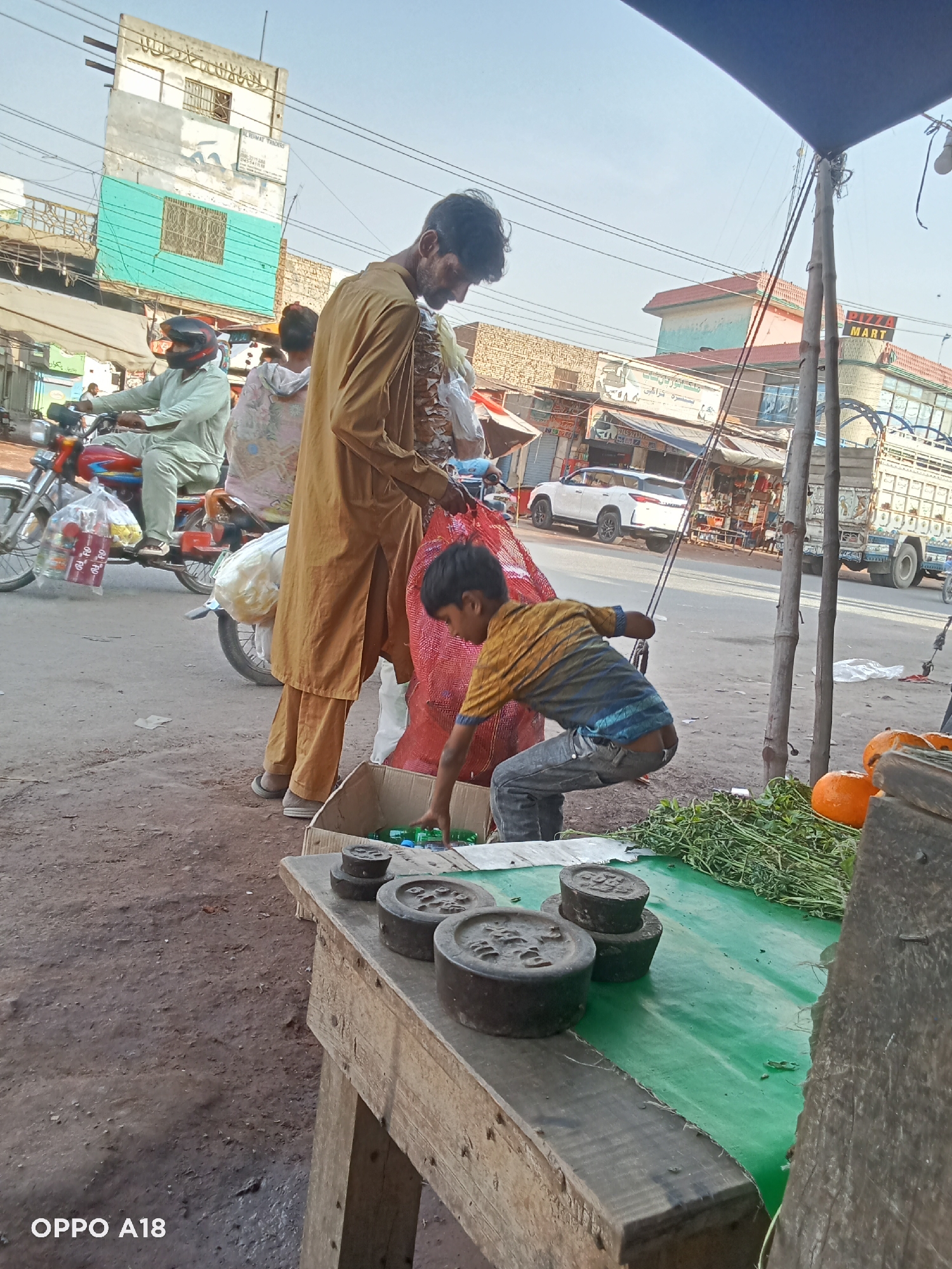 A medium, eye-level shot depicts a street scene in an urban environment.
In the foreground, a weathered wooden table covered with a green tarp displays various items. Several stacked, black, circular weights are visible towards the front left. Fresh, green leafy vegetables and a few orange pumpkins are on the table's right side. A cardboard box is below the table.
A man wearing a tan outfit stands in the middle of the frame, seemingly sorting through items. A child in a striped shirt and jeans bends over, reaching into the cardboard box.
In the background, a man in a light-colored outfit is riding a red motorcycle with bags hanging on the back. Various buildings, shops, and vehicles line the street. Signs in an unfamiliar script are visible on the buildings. Overhead wires crisscross the sky.
The overall scene suggests an outdoor market or street vendor setting.