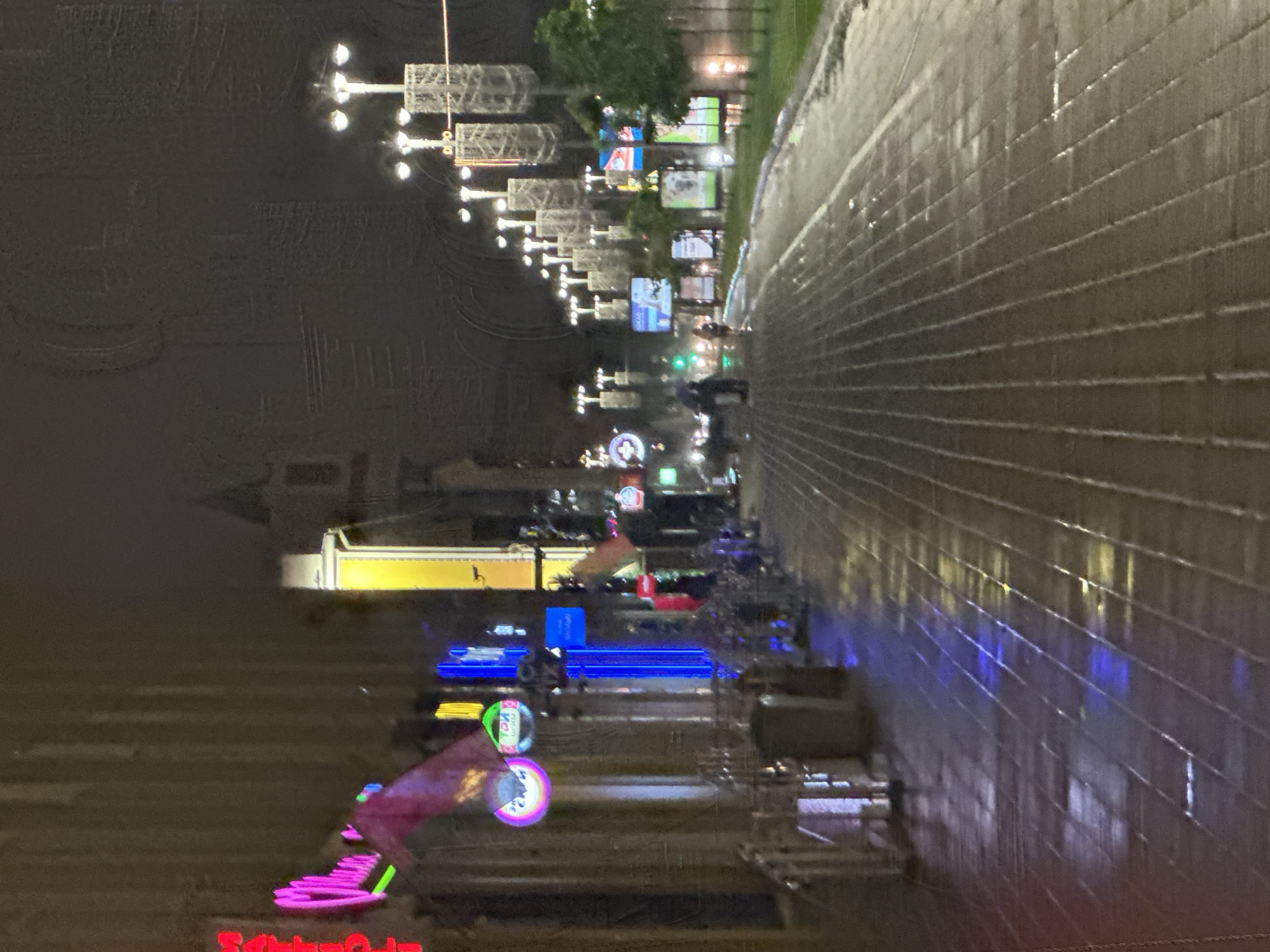 The image depicts a nighttime scene on a city street in Minsk, Belarus. The street is paved with cobblestones and appears wet, reflecting the colorful neon lights from buildings lining the left side of the street. These lights come from signs of businesses, including what appears to be a restaurant or bar with a prominent pink neon sign. The right side of the street features streetlights and large digital billboards displaying various advertisements. Several trees are visible on the right, and some pedestrians can be seen walking on the wet pavement. The overall atmosphere is that of a lively, illuminated urban environment at night.
