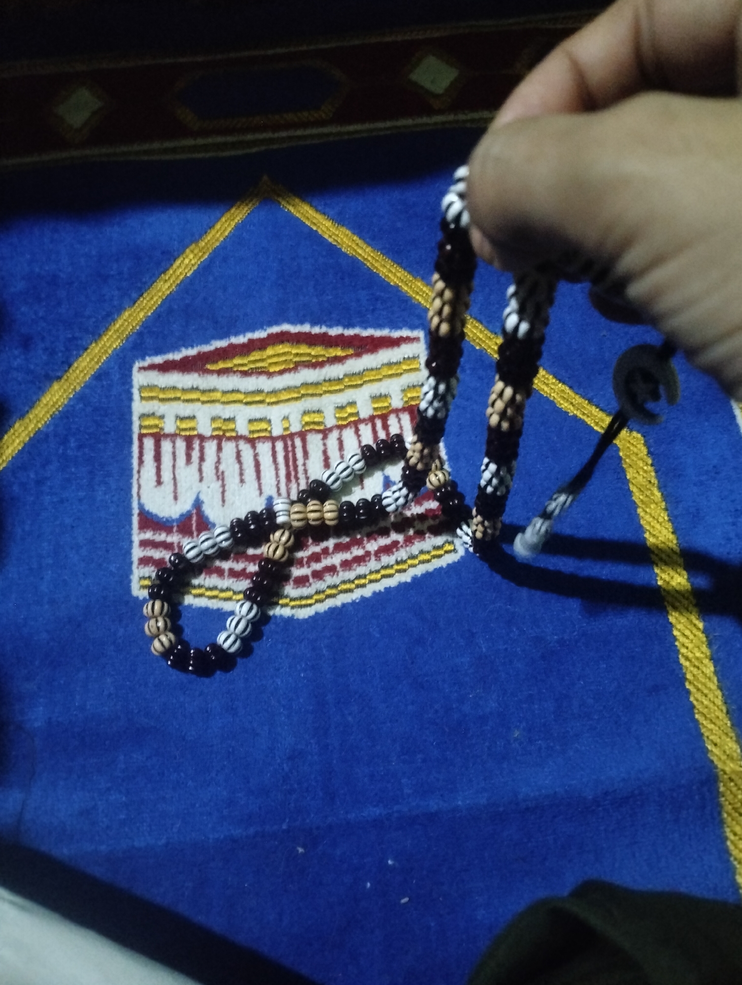 A hand is holding a string of prayer beads, which are made of alternating dark brown, light tan, and white beads. The beads are arranged in a circular pattern. The prayer beads are resting on a blue mat with a yellow border, which has an embroidered image of the Kaaba in the center. The mat is likely a prayer mat, commonly used by Muslims for prayer. The image was taken in Jetis, Indonesia.