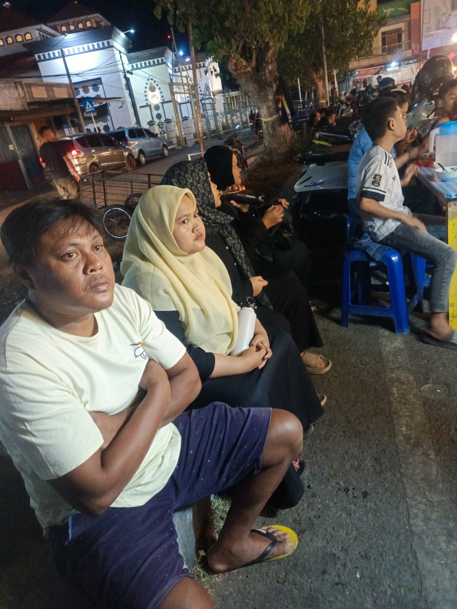The image depicts a street scene at night, likely in Luwoo, Indonesia, with several people gathered. In the foreground, a man wearing a light yellow t-shirt and dark blue patterned shorts sits with his arms crossed, looking off to the right. Beside him, a woman in a yellow hijab and a dark patterned garment is also seated. Further back, more individuals are visible, some seated at tables, suggesting a food stall or dining area. A mosque with white architecture and arched windows is visible in the background, illuminated by lights. Several vehicles, including cars and motorcycles, are parked along the street. The overall environment appears to be a bustling urban night setting with artificial lighting casting shadows.