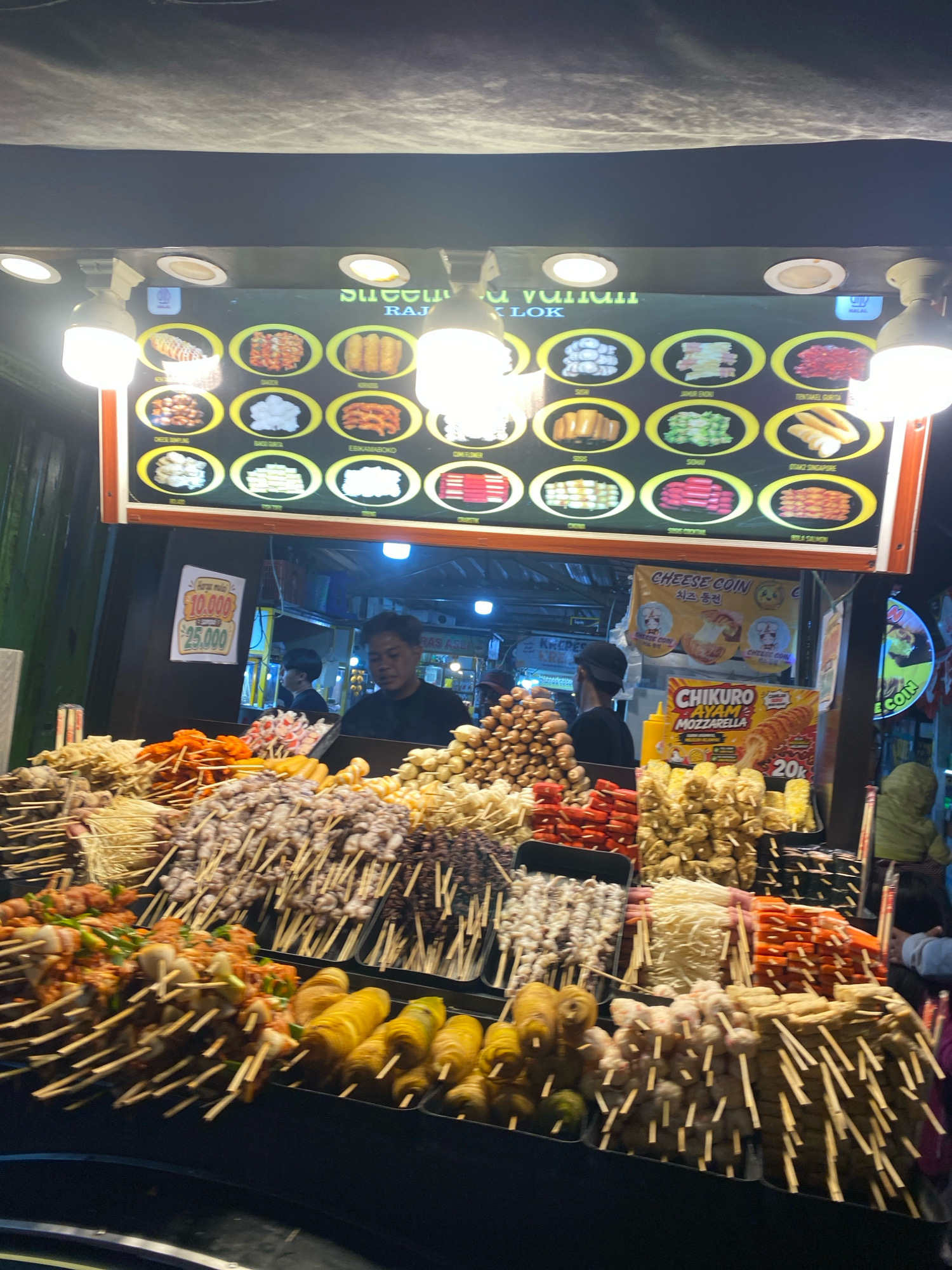 The image shows a busy street food stall at night in an unknown city. The stall is brightly lit and displays a wide variety of skewers of food, including what appear to be seafood, meat, and vegetables. A menu board above the stall shows pictures and prices of different food items, with some text in what appears to be Indonesian. Several people are visible in the background, including vendors and customers. The overall scene suggests a lively and bustling atmosphere typical of a night market.