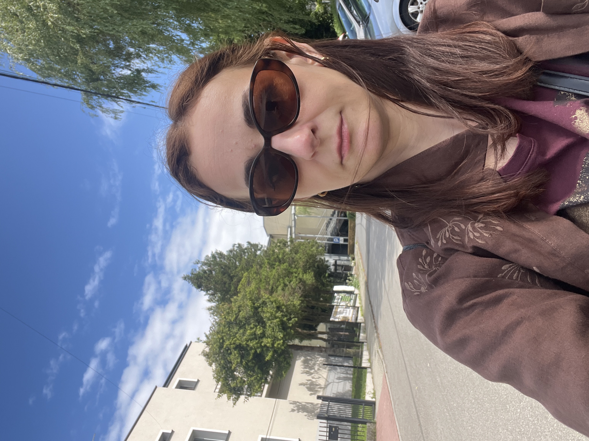 A tilted selfie shows an adult female outdoors on a sunny day. She has long brown hair and wears large, oval-shaped brown sunglasses, which obscure her eyes. Her lips are closed, suggesting a neutral expression. She is dressed in a brown jacket or shirt, possibly linen, featuring subtle embroidered floral or leafy patterns on the shoulders and sleeves. Underneath, a maroon-colored top with a shiny, possibly metallic, design is visible.
The background features a vibrant blue sky with scattered white clouds, indicative of clear weather. Lush green tree foliage fills parts of the frame, with some trees directly behind the subject and others further in the mid-ground next to a building. A light-colored, possibly beige or off-white, multi-story building with visible windows is situated in the lower-left. A paved street or sidewalk is seen in the foreground and middle ground, with a dark metal fence running along it. A silver or white car is partially visible in the upper-right background. Thin power lines traverse the sky. The scene suggests a casual daytime outing in an urban or suburban environment. No discernible text is visible anywhere in the image.