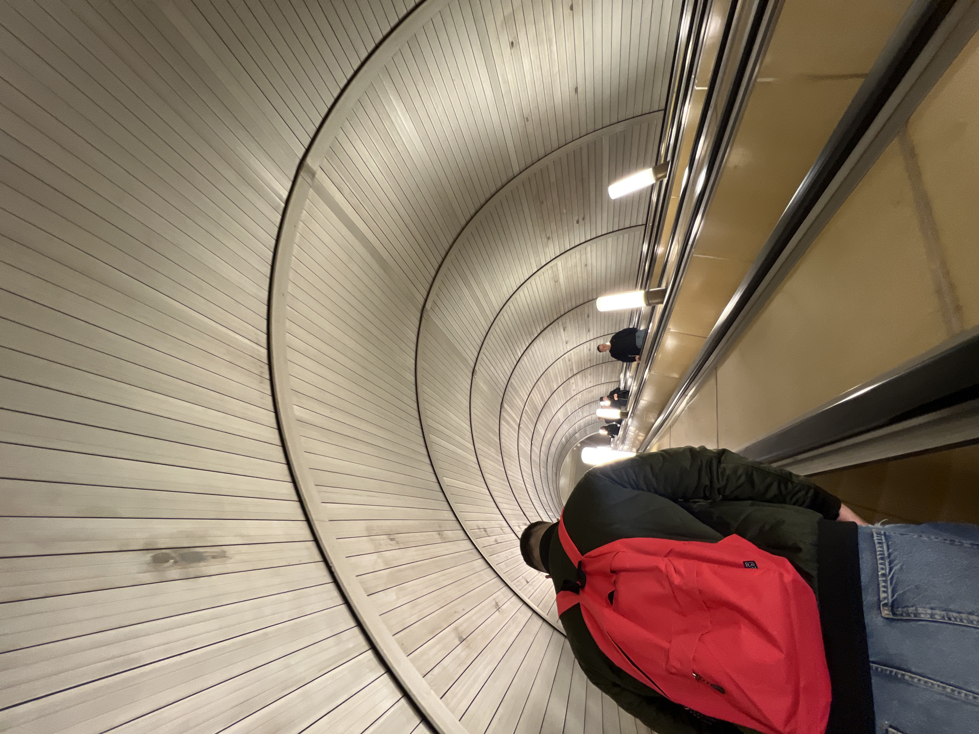 The image captures an unusual perspective inside a tunnel or passageway, likely an underground transportation hub. The scene is dominated by a curved wall made of light-colored, horizontally arranged wooden planks. The curvature creates a sense of depth and draws the viewer's eye into the distance. On the right side of the frame, there is an escalator with passengers on it. A person wearing a green jacket and blue jeans, and carrying a red backpack is visible in the foreground. Several other people are on the escalator in front of him, but they are further away. The tunnel is illuminated by regularly spaced light fixtures. The location is Park Pobedy Metro station in Moscow, Russia.