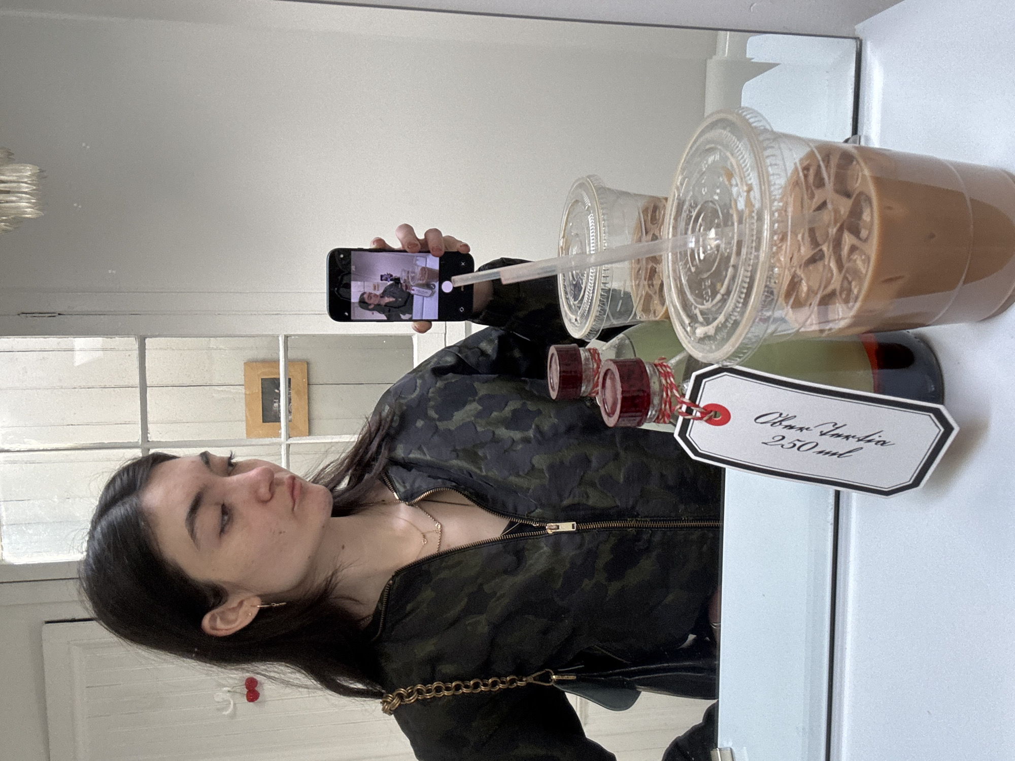 The photograph captures a woman taking a photo of a mirror reflection. In the reflection, the woman holds up her smartphone to photograph the image of herself holding her smartphone, in the context of a cold beverage setup. The cold beverage setup includes several bottled concoctions of some kind with labels and a tag, plus plastic cups filled with ice and cold coffee or tea. The woman is wearing a patterned jacket and a necklace. The location appears to be indoors, possibly in a café or shop in New York City, United States of America. The environment includes elements like a white-painted door or wall and a window.