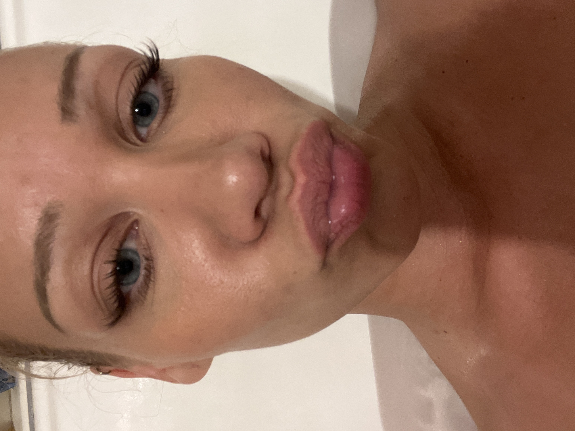 A close-up, low-angle, over-the-shoulder shot of a woman in a bathtub is presented. The woman's face fills the majority of the frame, with her eyes closed and lips pursed in a kissing gesture. She has blue eyes, dark eyelashes, and light brown hair pulled back. The water of the bathtub is visible around her shoulders and neck. The background is a plain white wall. The image does not contain enough information to determine its location in Mühldorf a. Inn, Germany.