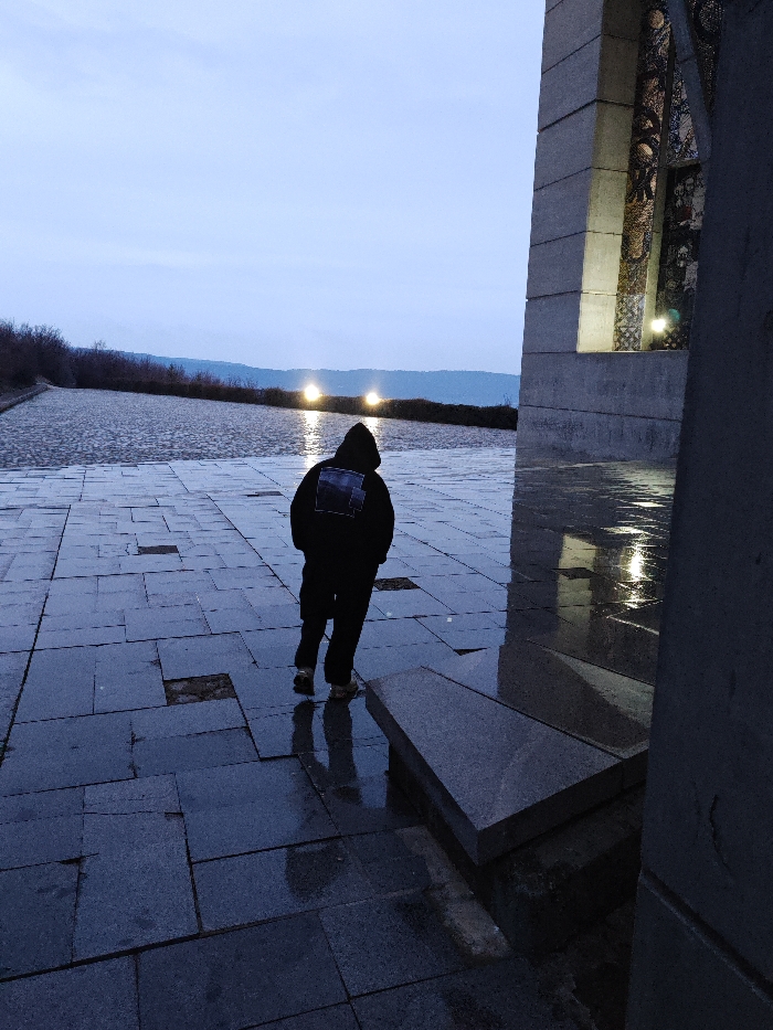 A single individual walks away from the camera across a wet, dark grey paved plaza at the Monument to 1300 Years of Bulgaria in Shumen, Bulgaria. The subject wears a dark, hooded jacket featuring a lighter-toned graphic design on the back, dark trousers, and light-colored shoes; their lower body reflects on the water-slicked flagstones. To the right, a large section of the monument's concrete architecture displays recessed panels containing illuminated mosaic artwork. Two prominent artificial lights within these recesses cast bright, elongated reflections on the wet pavement. Beyond the plaza, a wide, shallow reflecting pool or water feature shows rippled surfaces reflecting multiple horizontal lines of distant artificial lights. The background consists of dark, undulating hills with sparse vegetation under a pale, overcast sky, indicative of dusk or early night. Several paving stones on the plaza are damaged or missing, exposing the underlying earth. A rectangular, dark grey stone slab or bench is situated on the wet pavement in the lower right foreground.