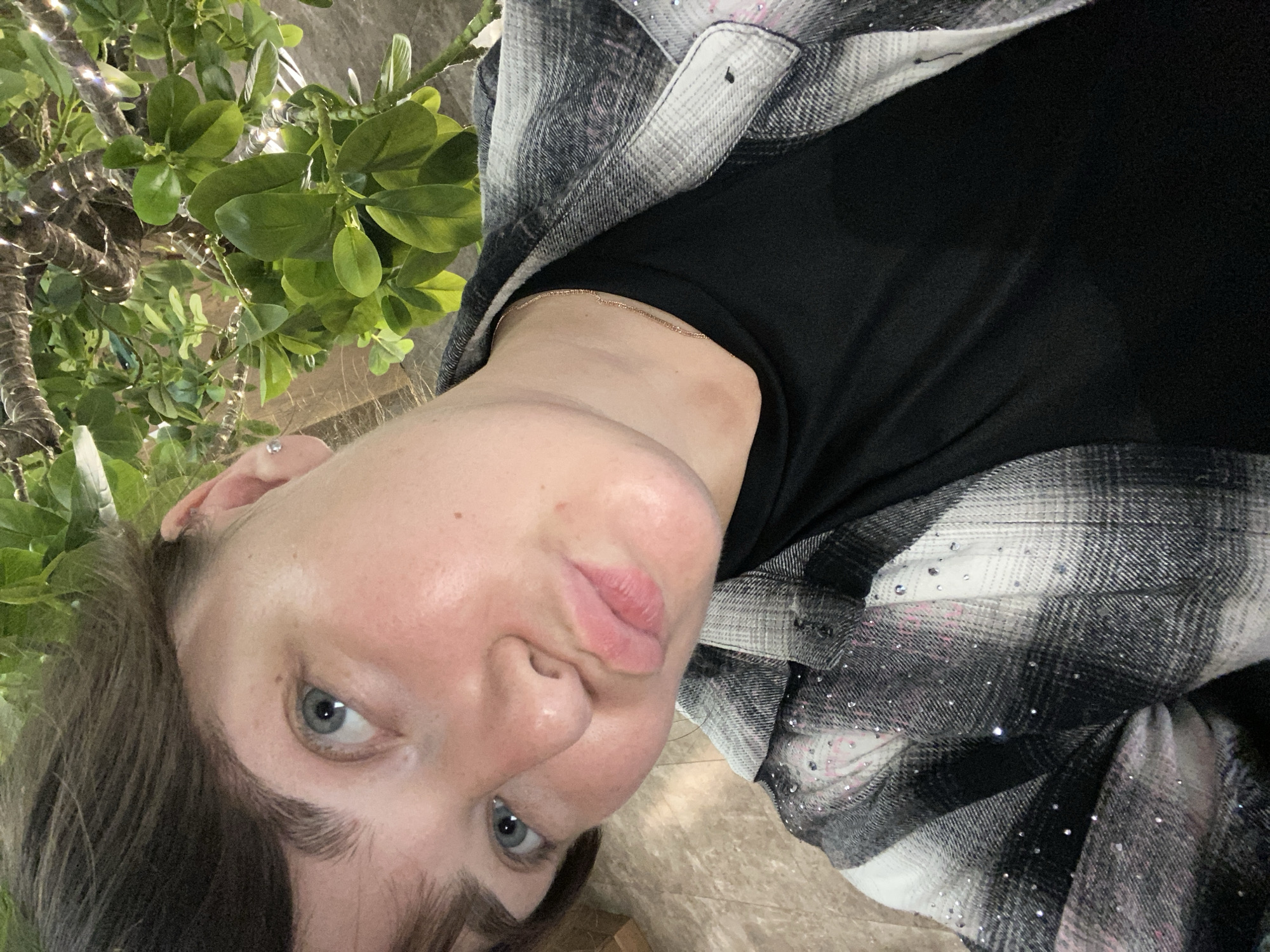 A young woman with short brown hair and blue eyes is pictured from the chest up, looking towards the camera with her lips pursed. She is wearing a black shirt and a gray and white plaid shirt adorned with rhinestones. The background consists of lush green leaves and branches, suggesting an outdoor setting, possibly a garden or park. The lighting suggests it is daytime. There are no recognizable landmarks or elements that would place the image specifically within Votkinsk, Russia.