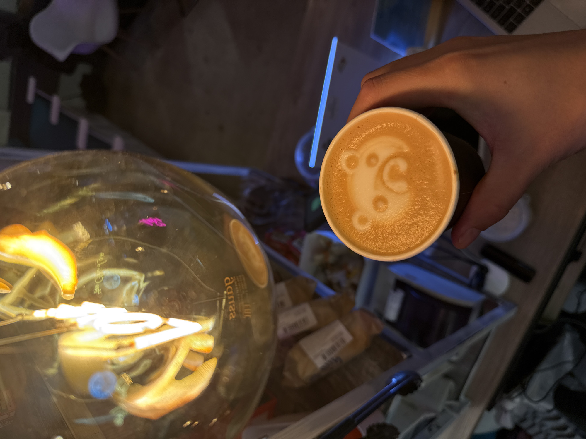 A close-up, over-the-shoulder shot shows a hand holding a coffee cup with a bear-shaped latte art design. The cup is dark brown and sits in the lower right quadrant of the frame. The background is blurred and dimly lit, but appears to be an indoor setting, possibly a cafe. In the upper portion of the image, a large, glowing light fixture with filament bulbs is visible, reflecting in a glass surface below. The overall impression is of a cozy, late-night atmosphere. There is no indication of the city of Moscow, Russia.